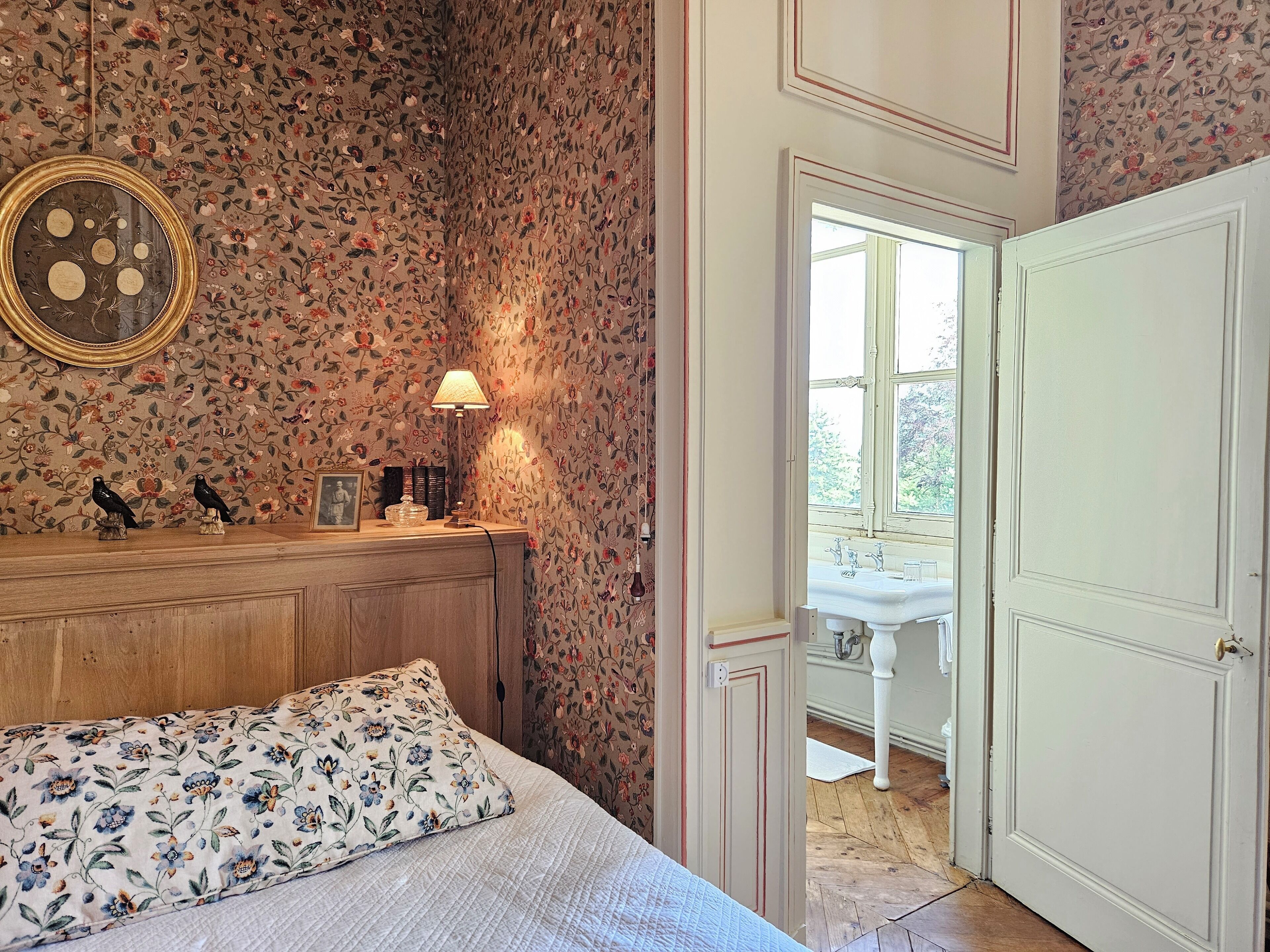 A bedroom with floral wallpaper, a wooden dresser, a bed with a floral pillow, a nightstand with a lamp, and a door leading to a bathroom with a sink and window.