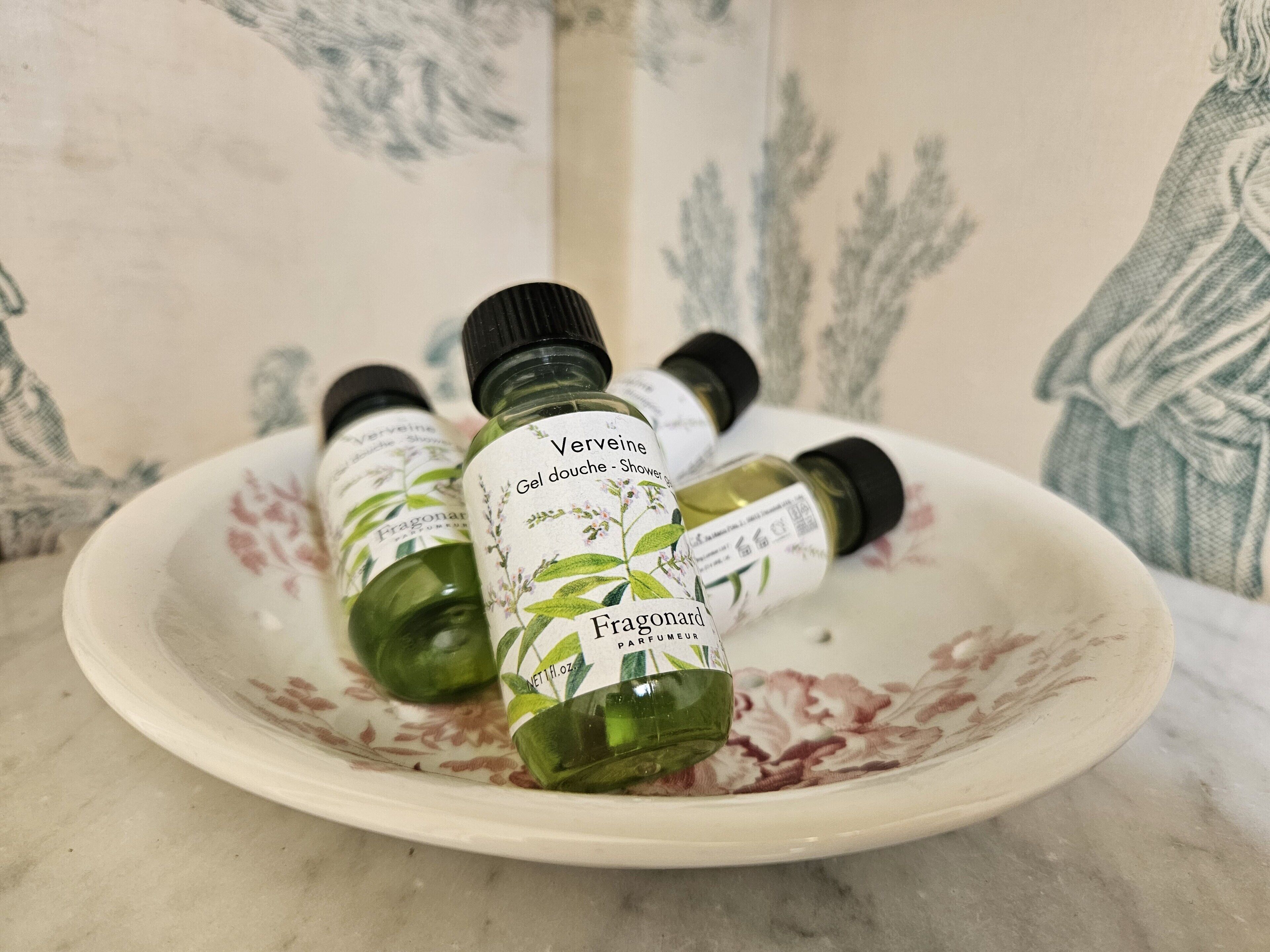 Un plato de cerámica blanca con cuatro botellas de vidrio verde de perfume Verveine Fragonard sobre una superficie de mármol.