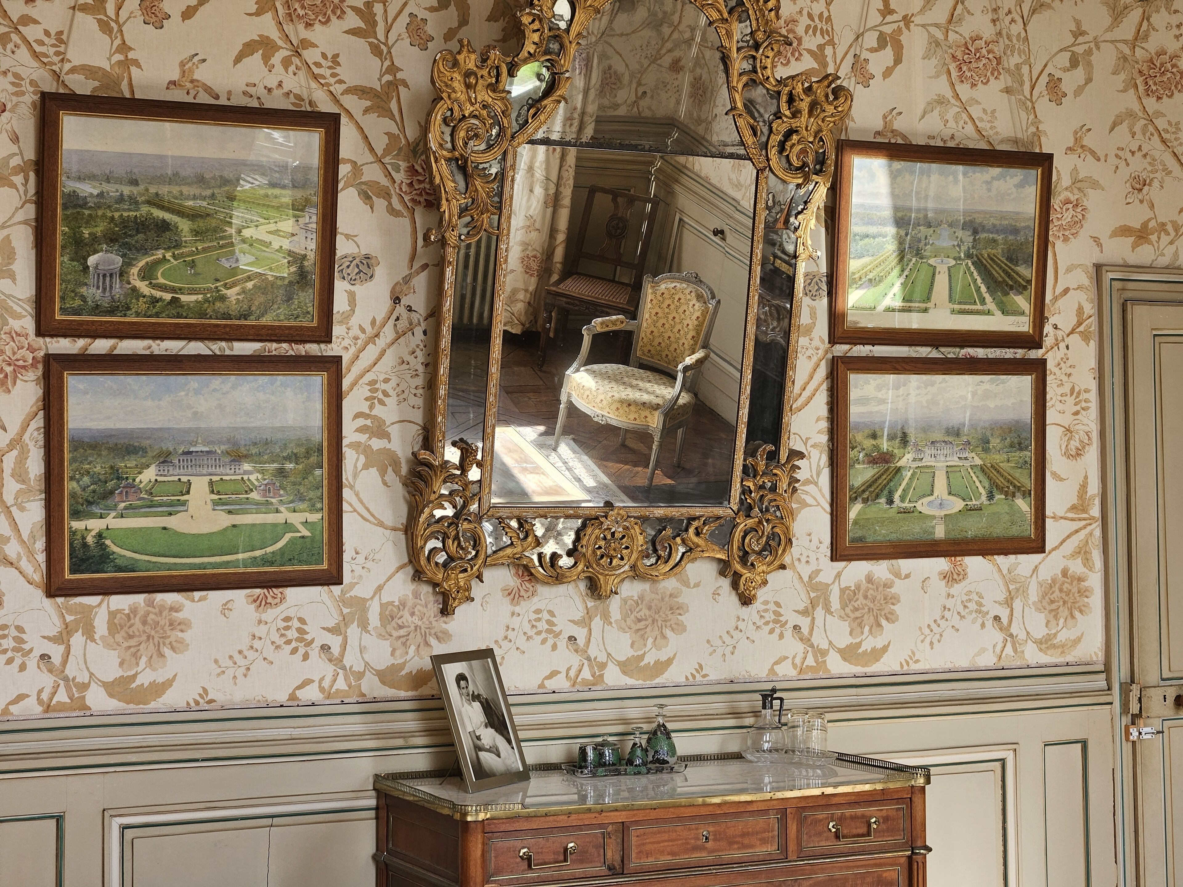 A room with a large ornate mirror, floral wallpaper, and framed landscape paintings.