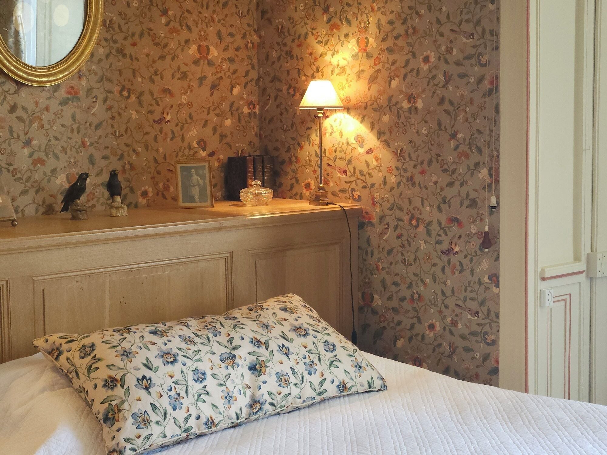 A bedroom with floral wallpaper, a bedside lamp, a wooden headboard, a bed with a floral pillow, and a small shelf with decorative items.