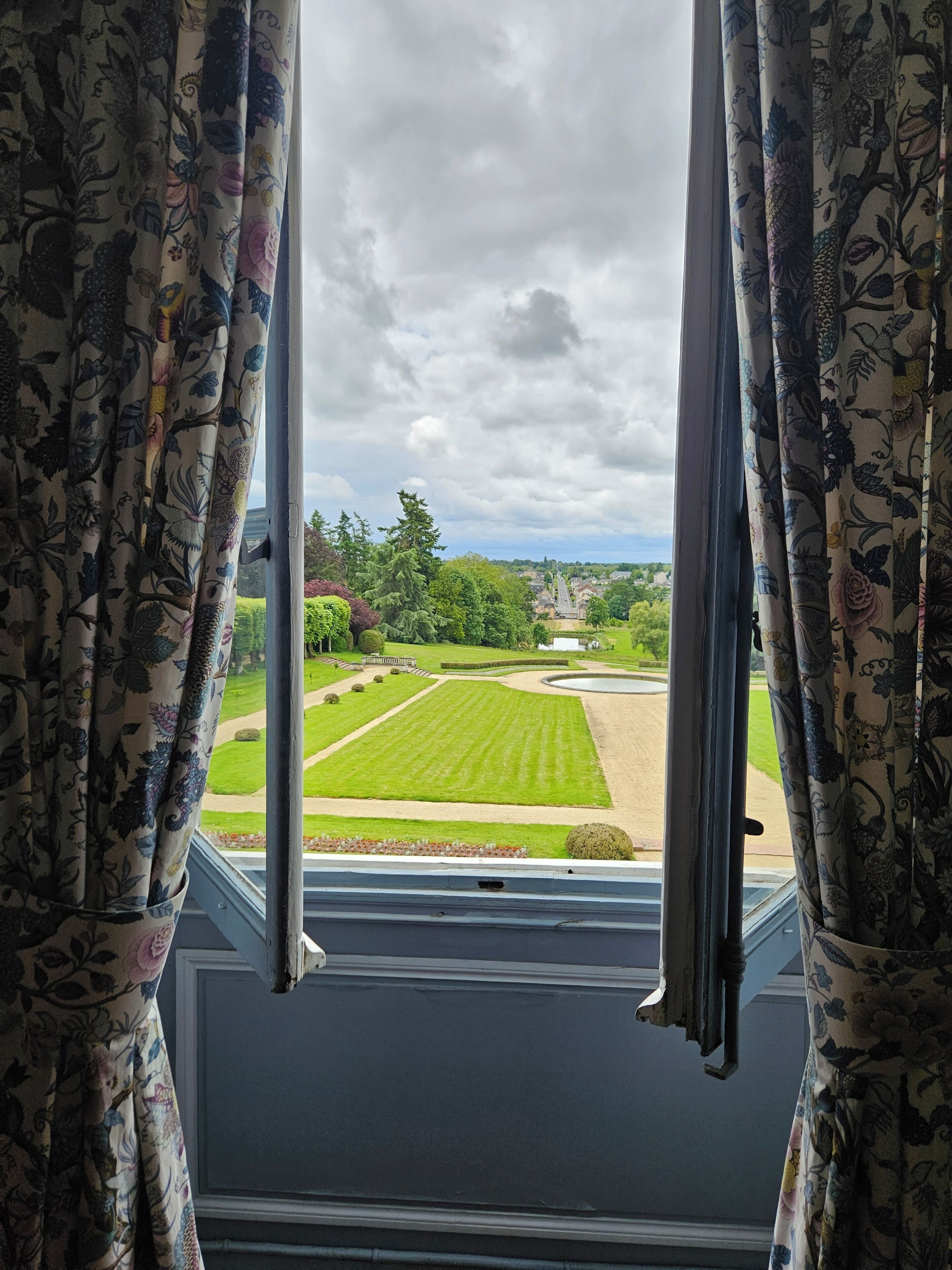 A view through a window with floral curtains, revealing a well-manicured lawn, a pathway, and distant buildings.