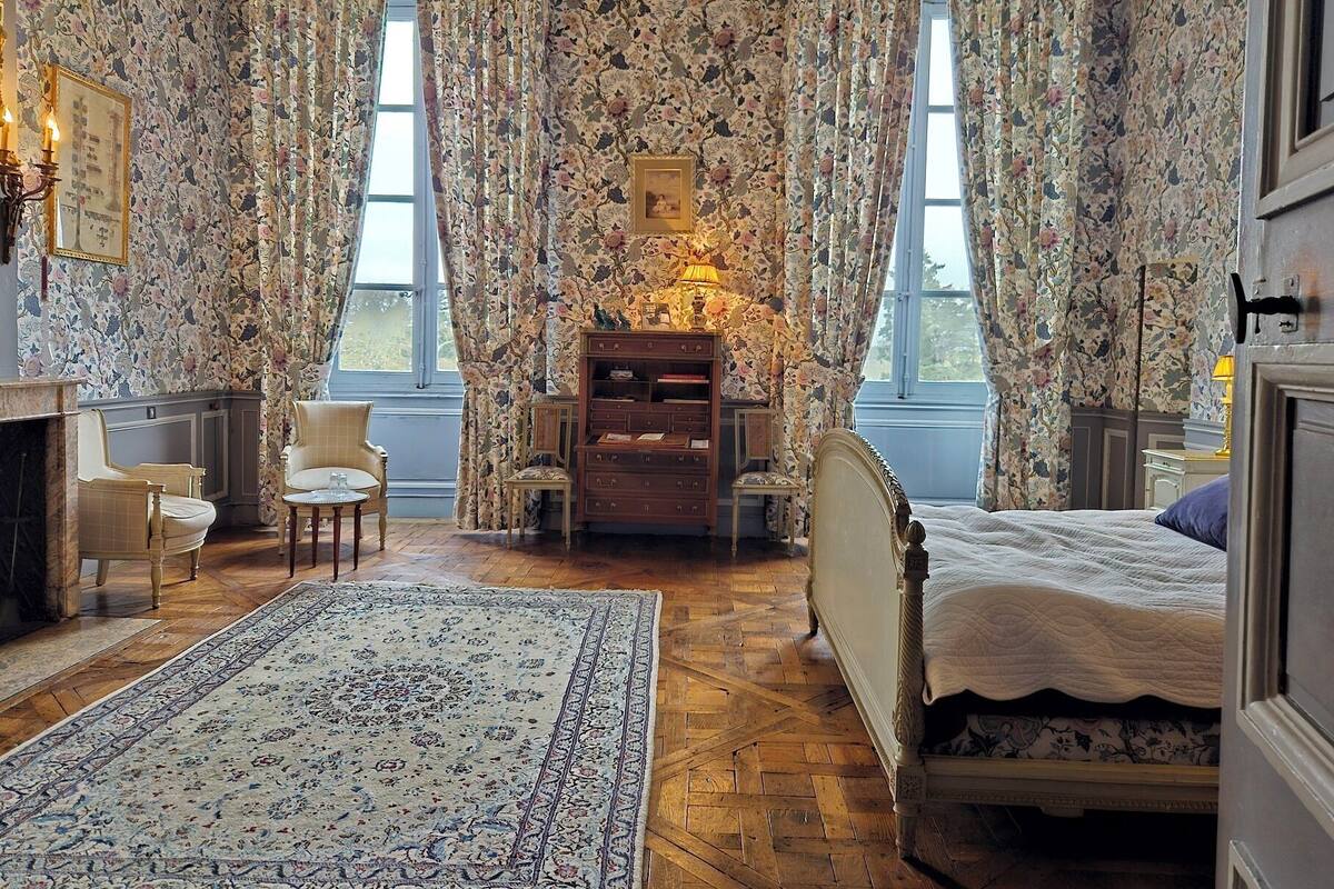 A room with floral wallpaper, a bed, a desk with a lamp, a chair, and a rug.