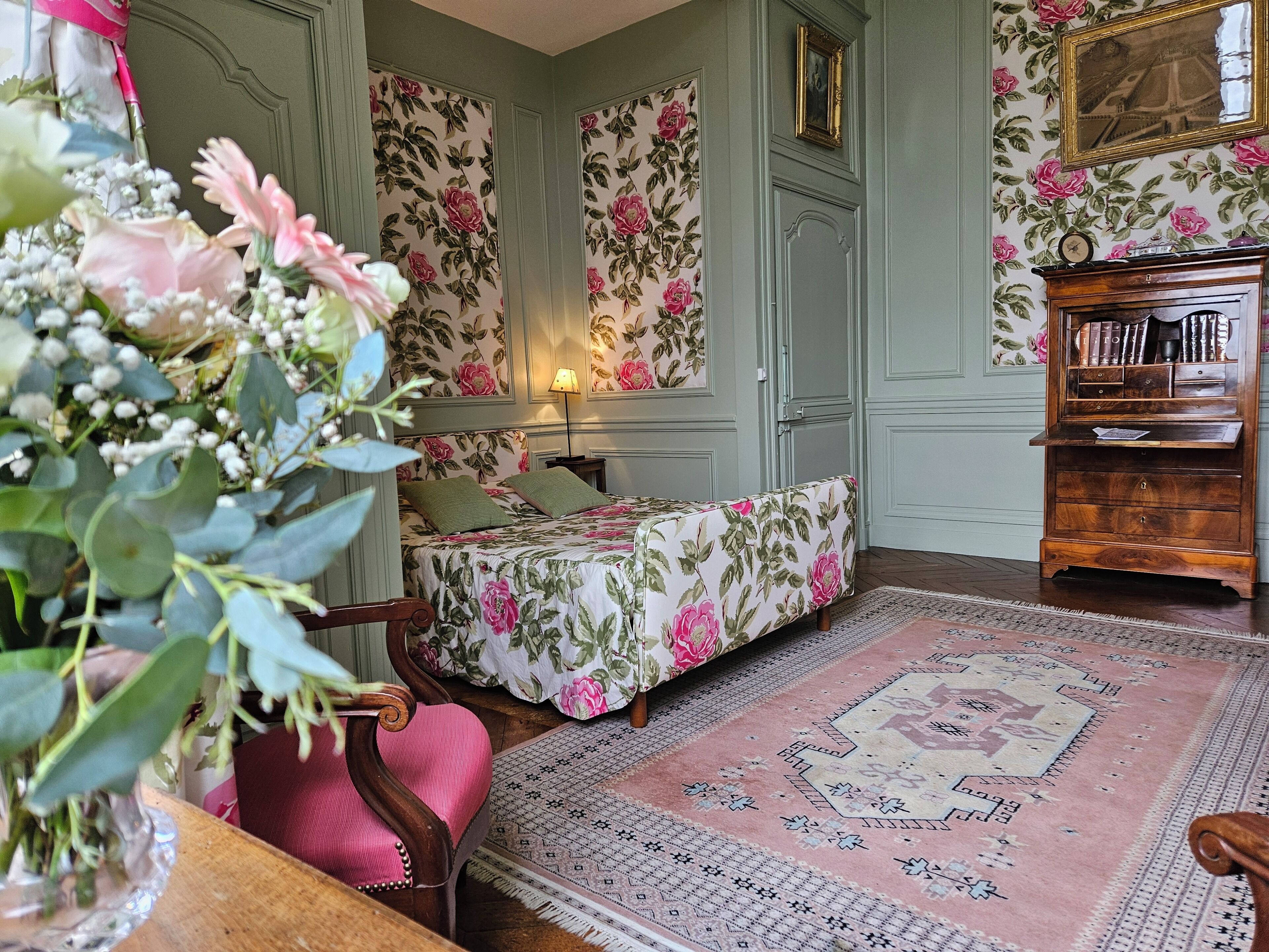 A room with floral wallpaper, a bed with green pillows, a wooden side table, a small lamp, and a bookshelf.