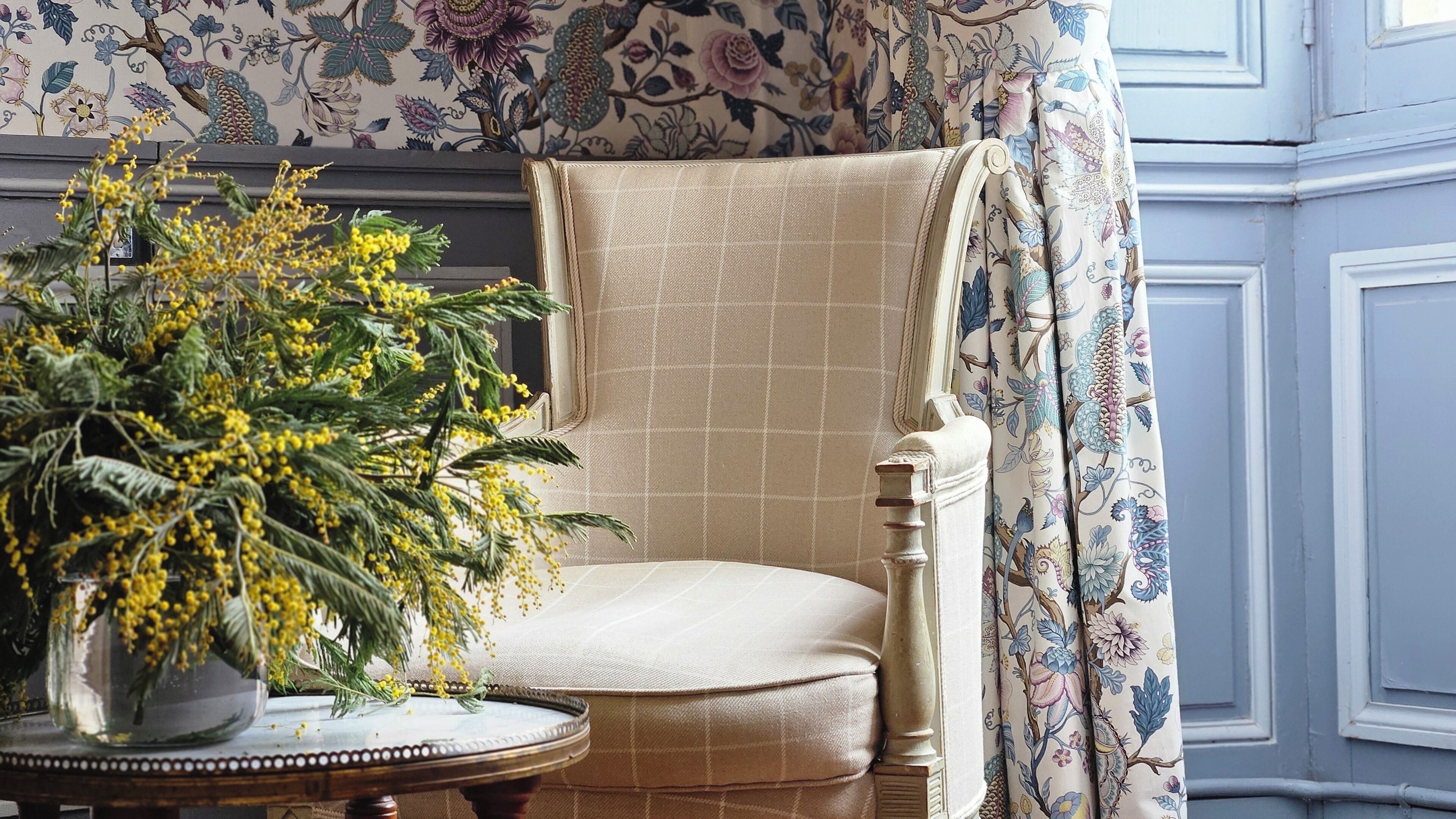 Ein Stuhl mit Karomuster, blumiger Tapete und eine Vase mit gelben Blumen.