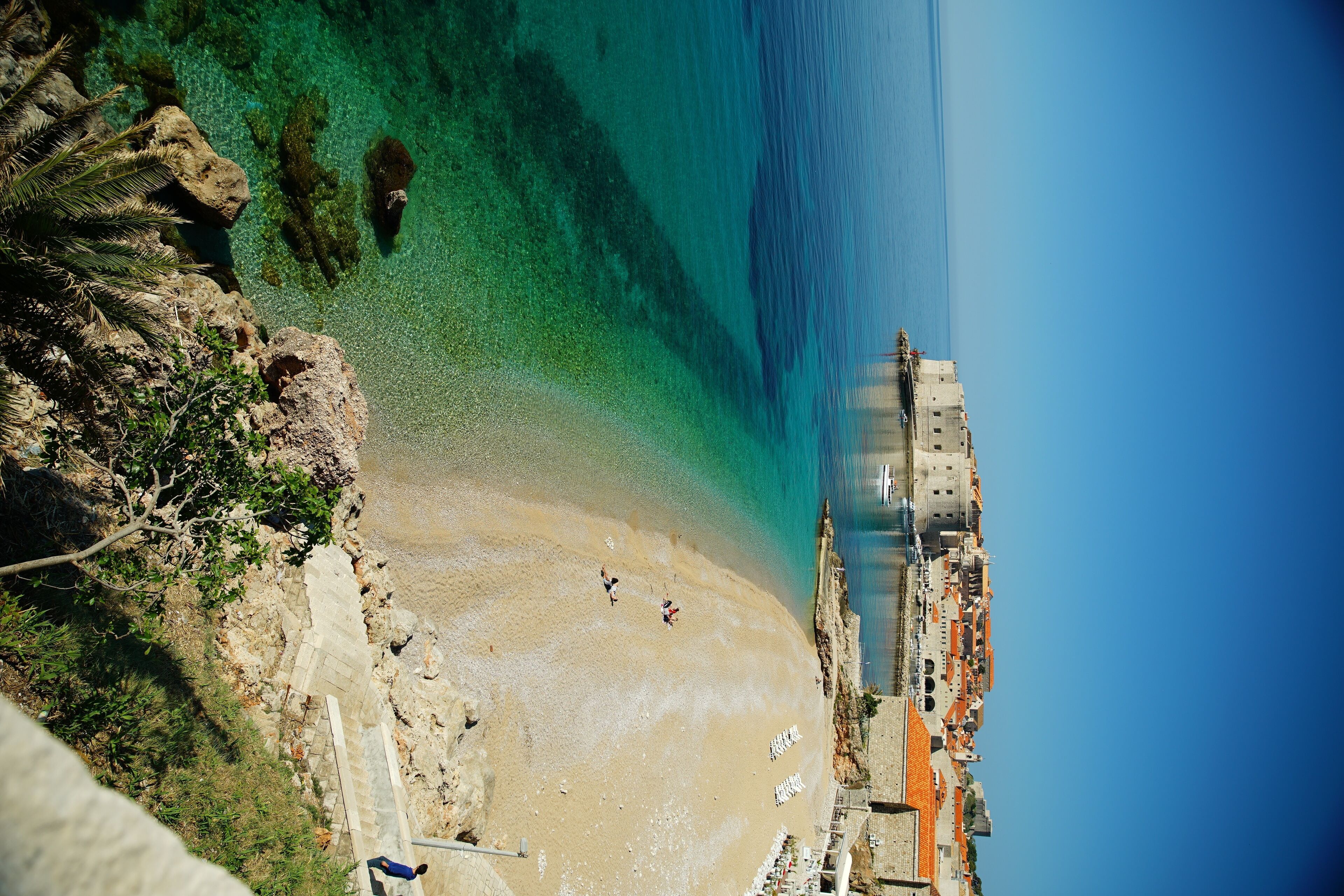 A coastal view with a beach, clear water, and a building with multiple levels and balconies.