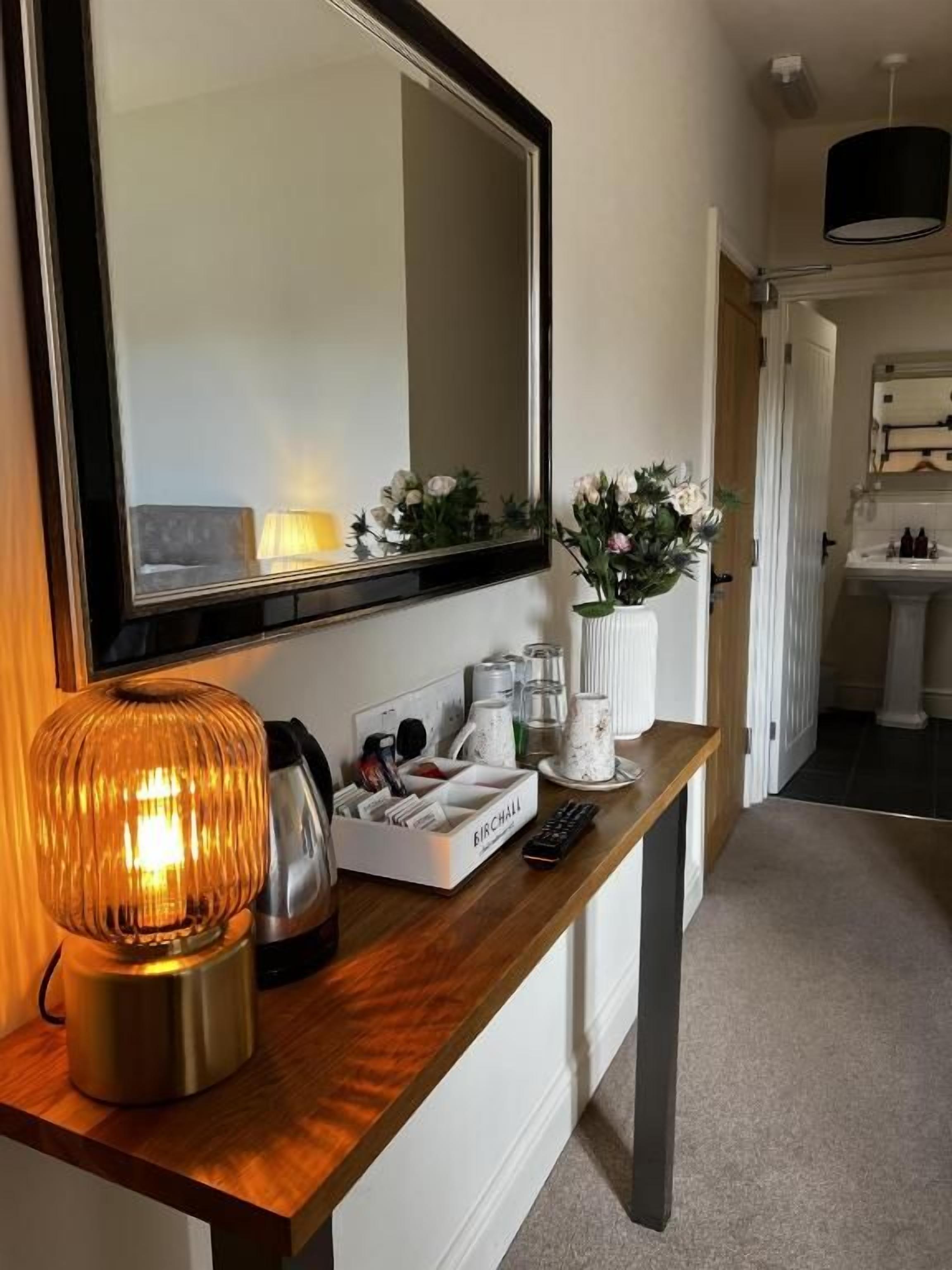 Ein Flur mit einem Holzkonsolentisch, einem Spiegel, einer Lampe, einer Vase mit Blumen und einem im Hintergrund sichtbaren Badezimmer.