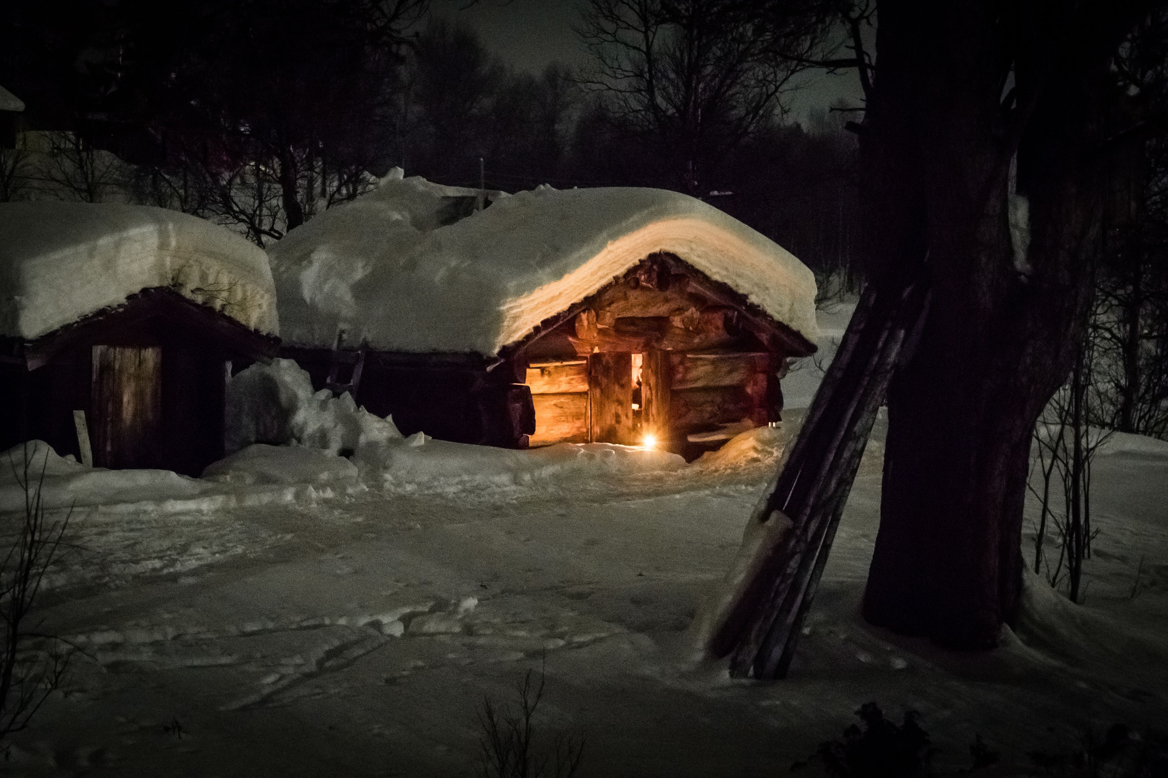 A snow-covered wooden cabin with a lit interior, surrounded by snow.