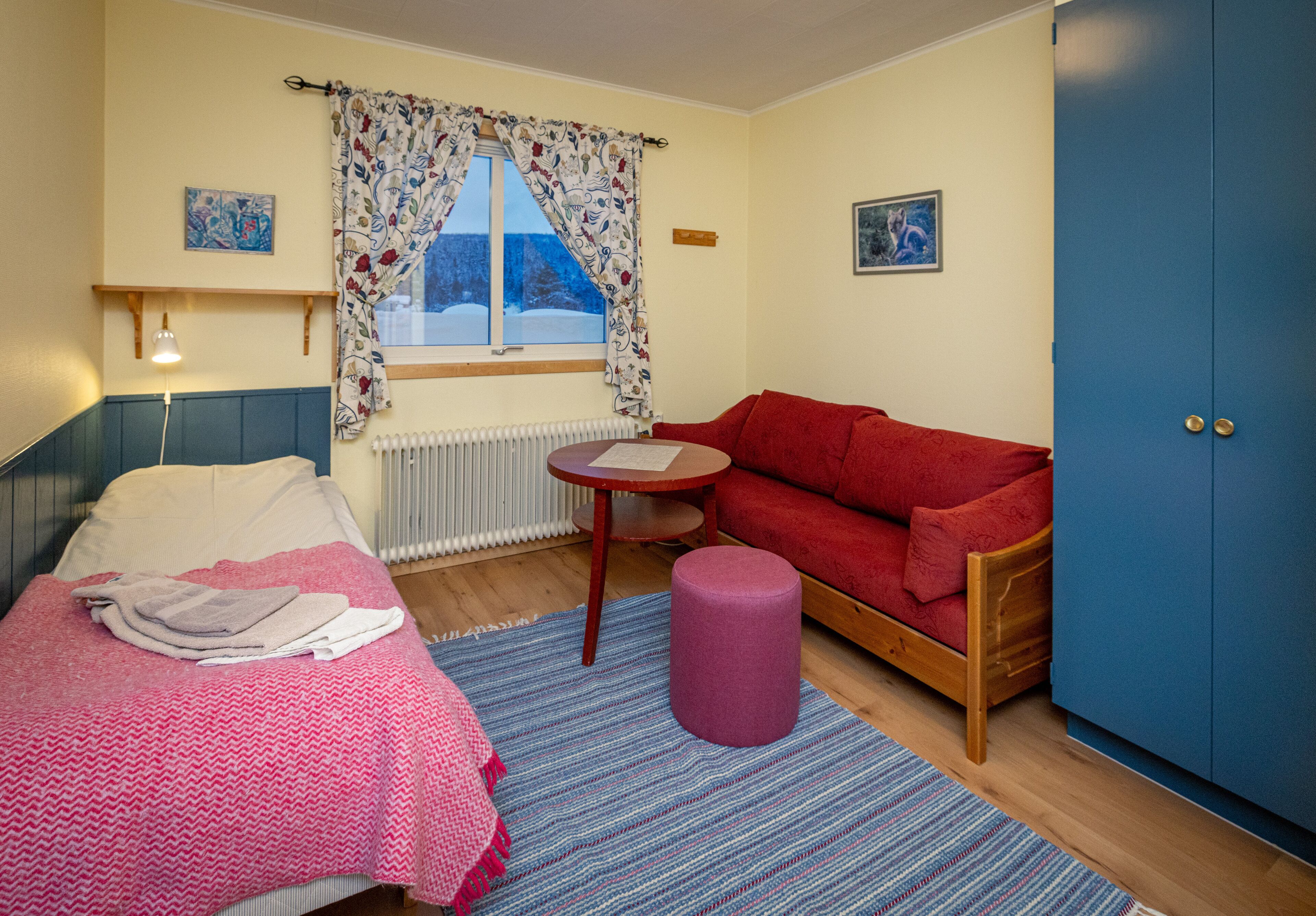 A room with a bed, a red sofa, a small round table, a purple ottoman, a window with floral curtains, and a blue wardrobe.