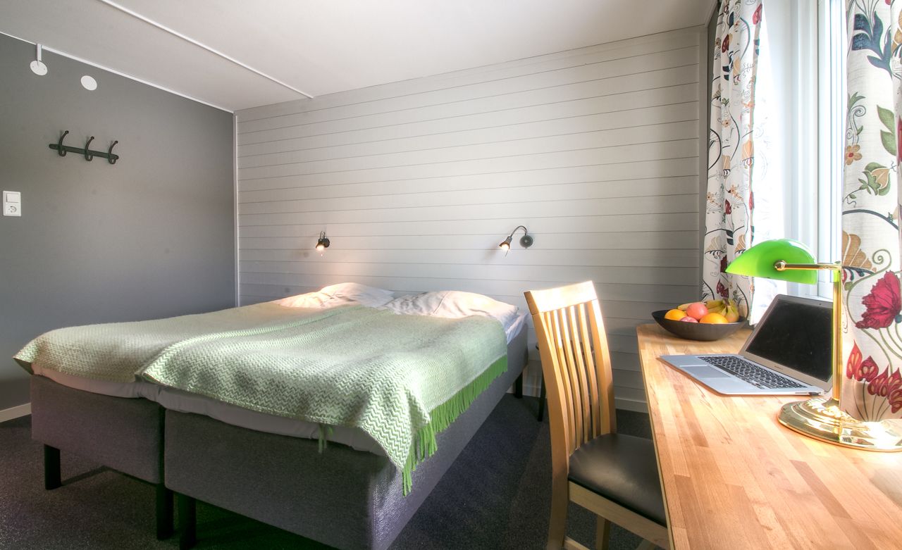 A bedroom with a bed, a desk with a laptop, a chair, a lamp, and a fruit bowl.