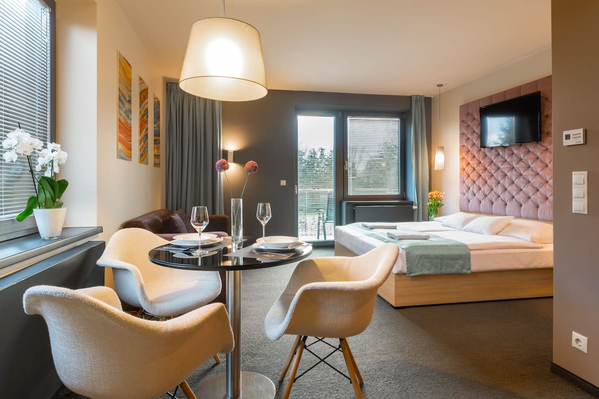 A modern hotel room with a bed, a dining table with chairs, a TV mounted on the wall, and artwork on the walls.