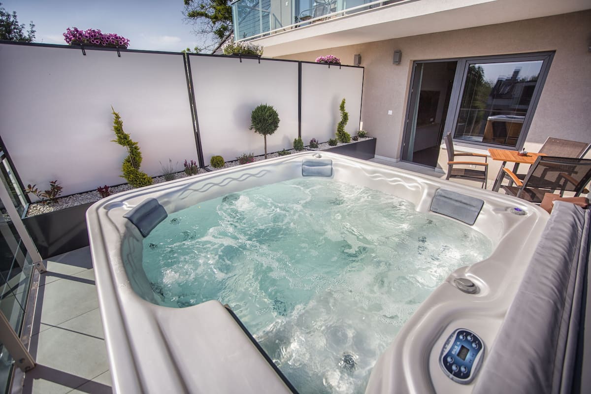A hot tub with a control panel, surrounded by a patio area with a table and chairs, and a view of a building in the background.