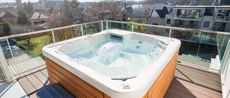 A hot tub on a wooden deck with a glass railing, overlooking a residential area.
