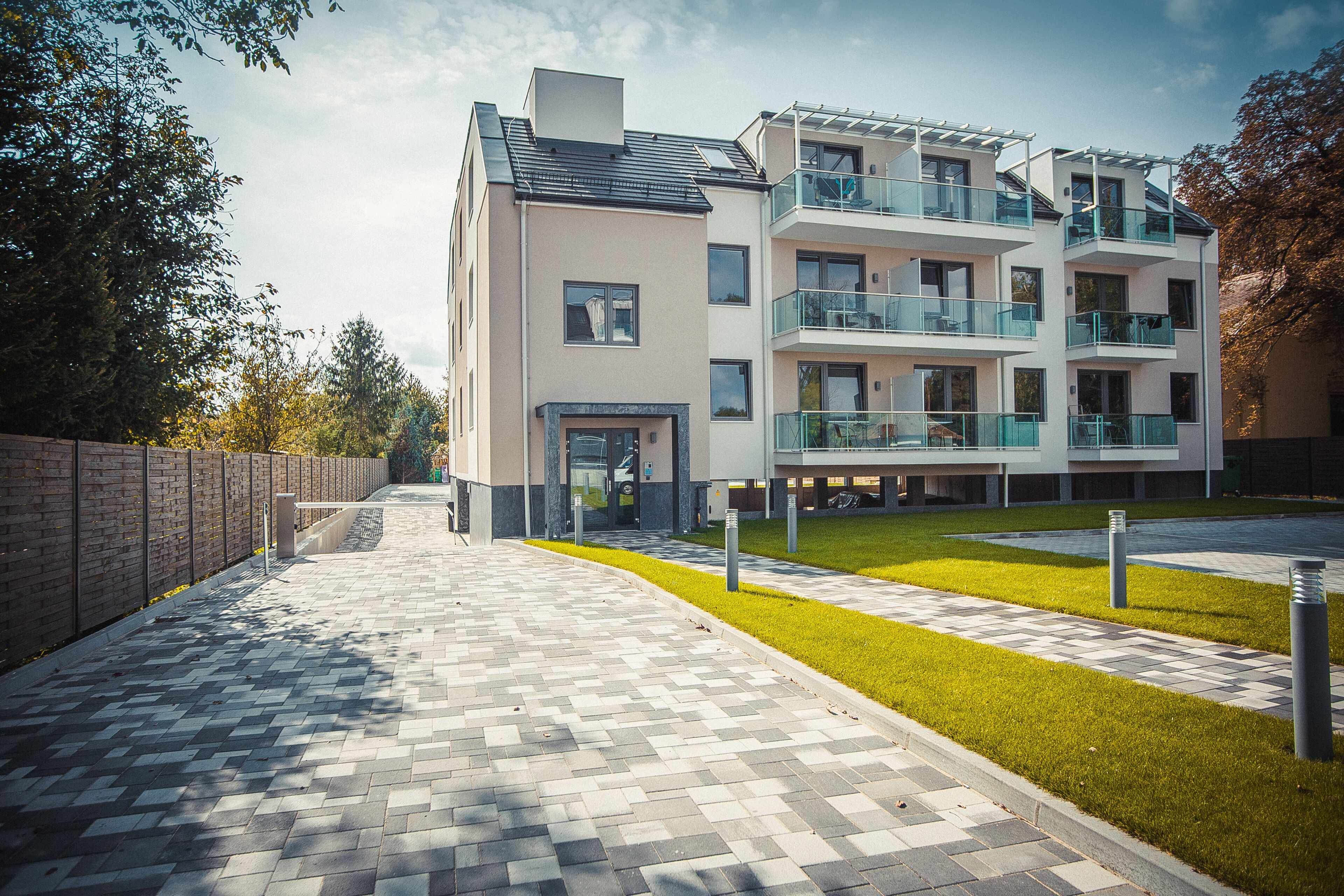 A modern multi-story residential building with a paved walkway, a grassy area, and a brick wall on one side.