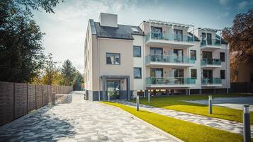 A modern multi-story residential building with a paved walkway, a grassy area, and a brick wall on one side.