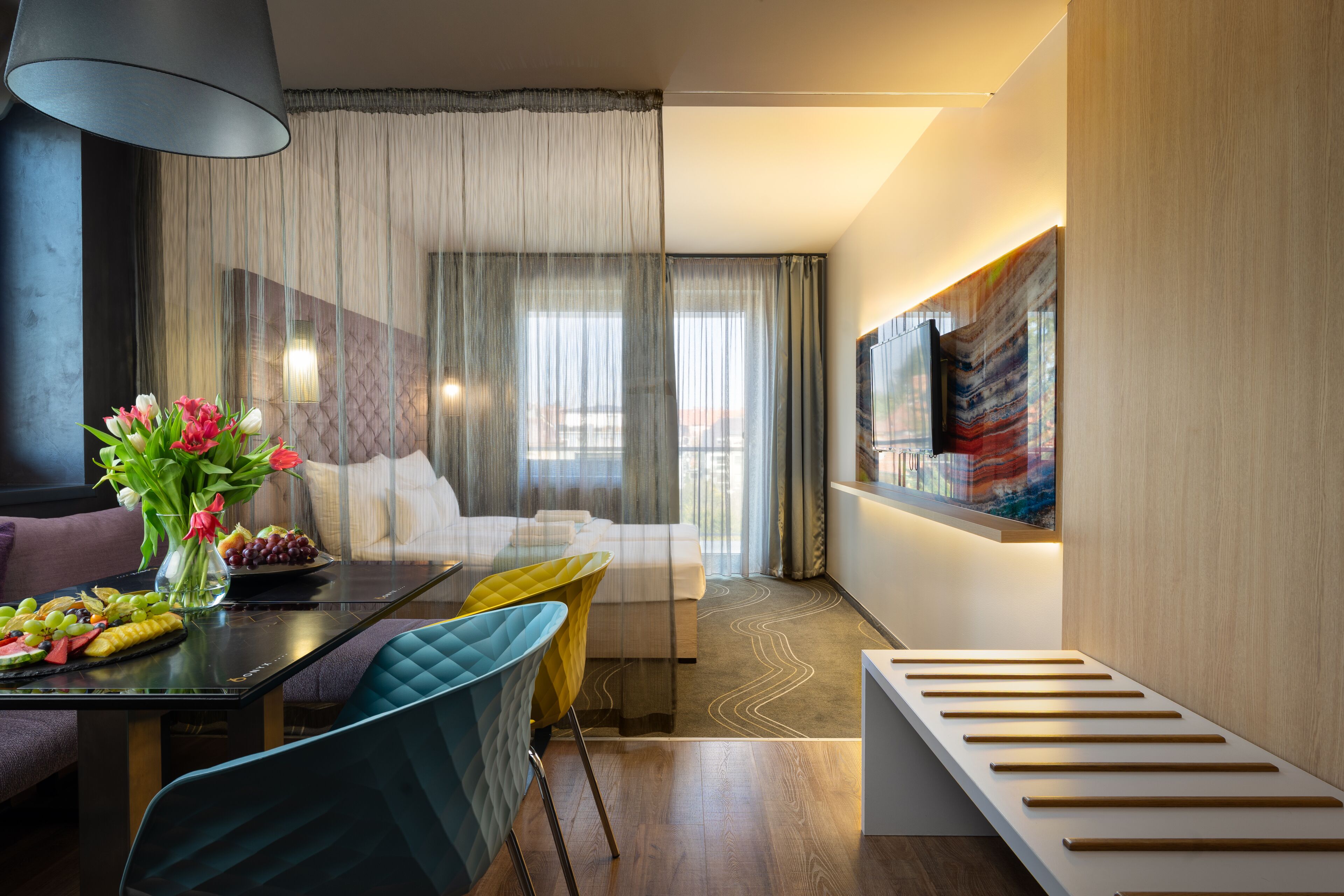 A modern hotel room with a bed, a dining table with fruit, a vase of flowers, and a large painting on the wall.