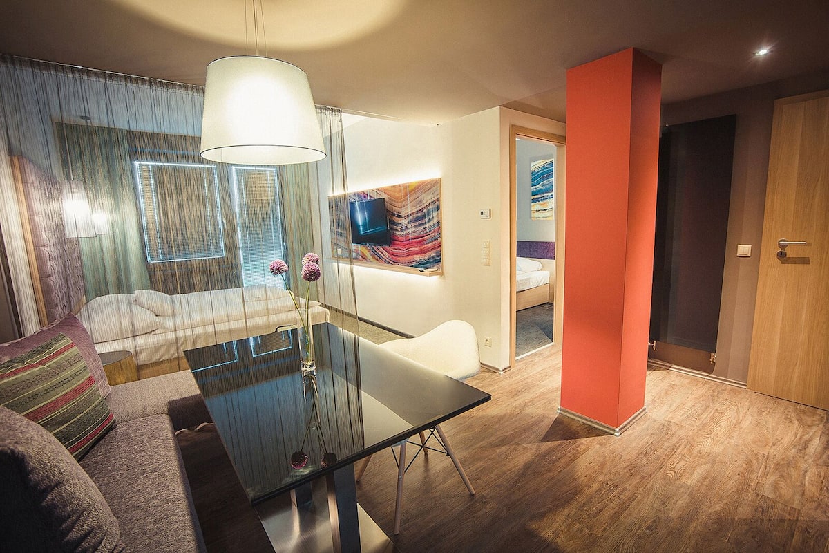 A modern hotel room with a glass dining table, a sofa, and a bed.