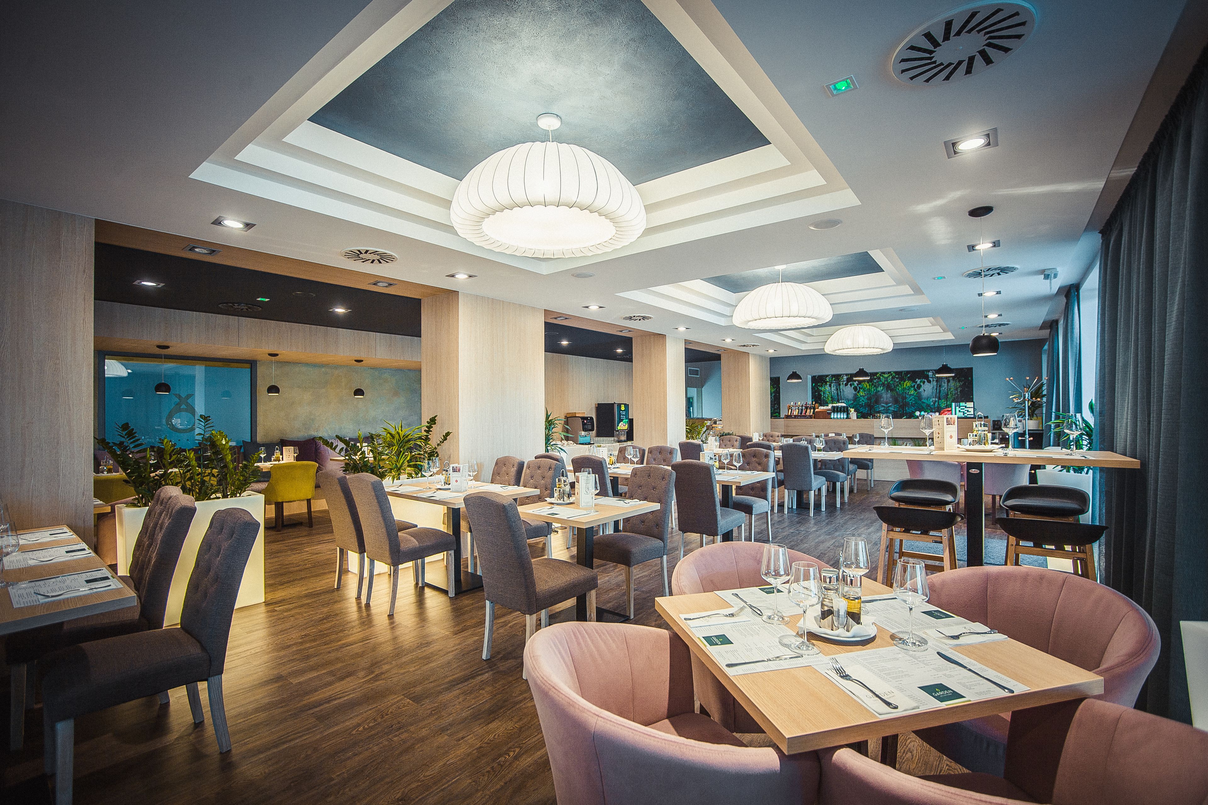 A modern restaurant interior with wooden flooring, multiple dining tables and chairs, pendant lighting, and a bar area.