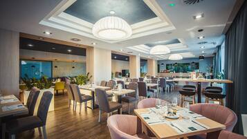A modern restaurant interior with wooden flooring, multiple dining tables and chairs, pendant lighting, and a bar area.