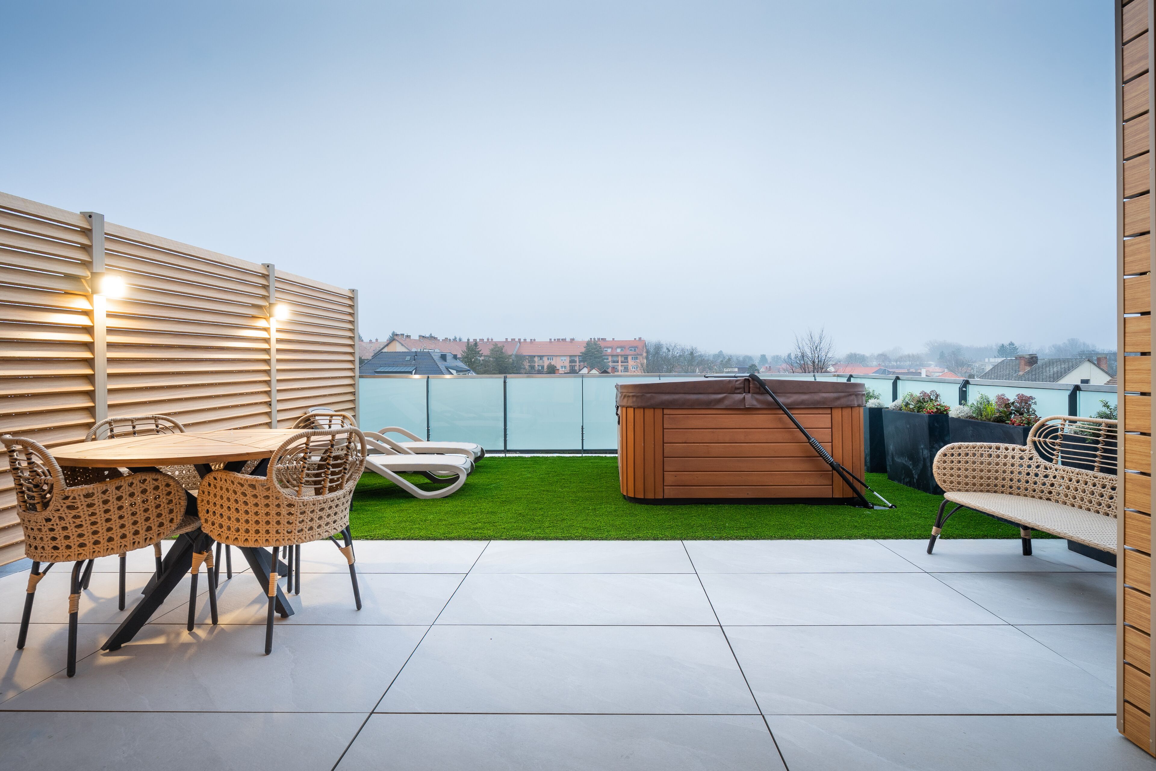 Un patio en la azotea con una mesa y sillas de madera, un jacuzzi y vistas a edificios en la distancia.