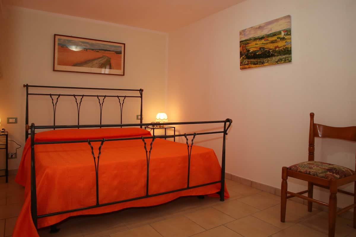 A bedroom with a metal bed frame, an orange bedspread, a wooden chair, a bedside table with a lamp, and two paintings on the walls.