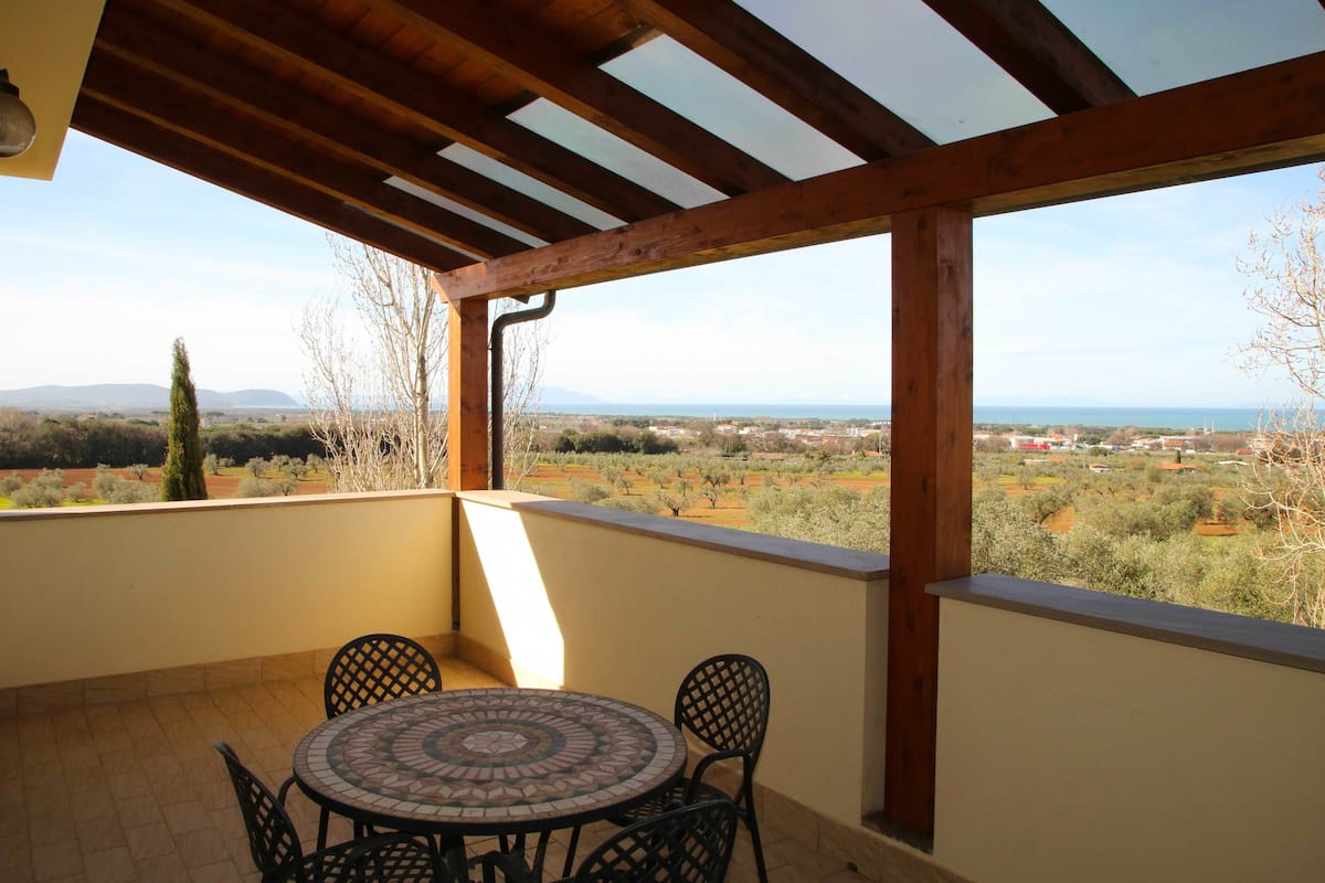 A covered patio with a table and chairs, offering a view of a rural landscape with trees and distant buildings.