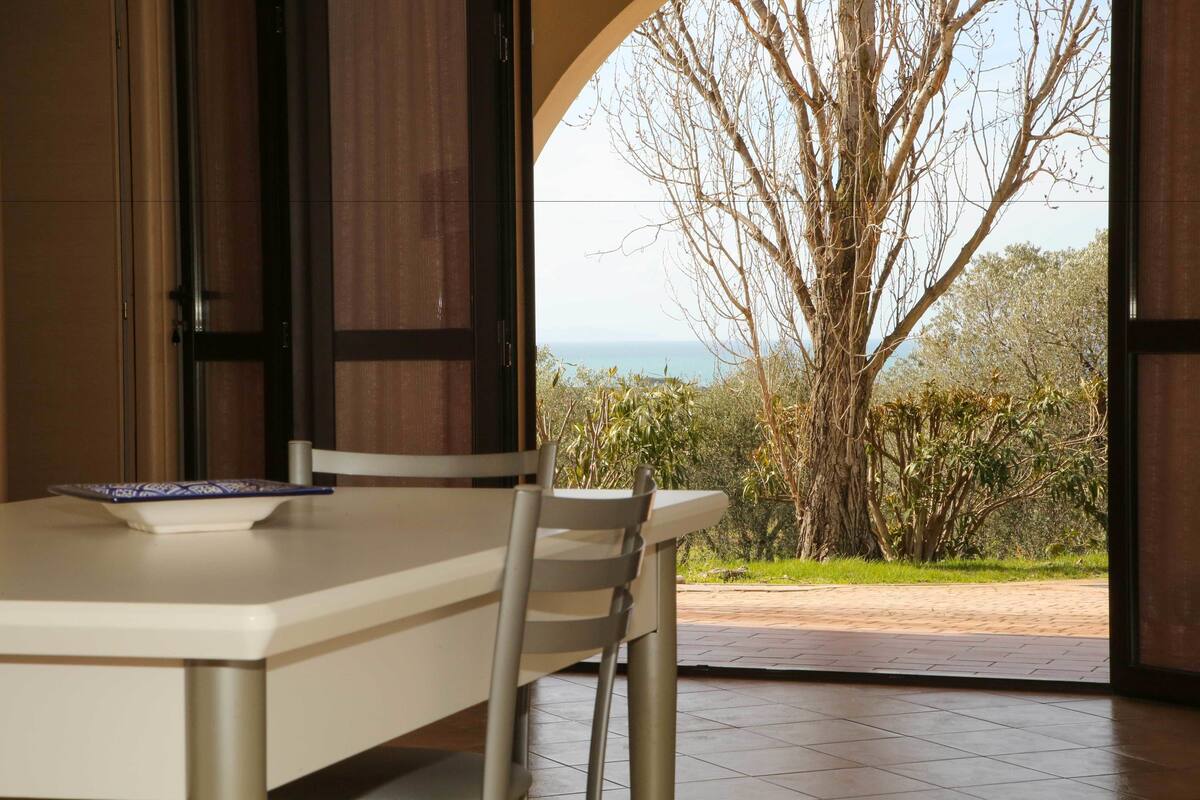 A dining area with a table and chairs, a view of the outdoors including trees and a body of water.