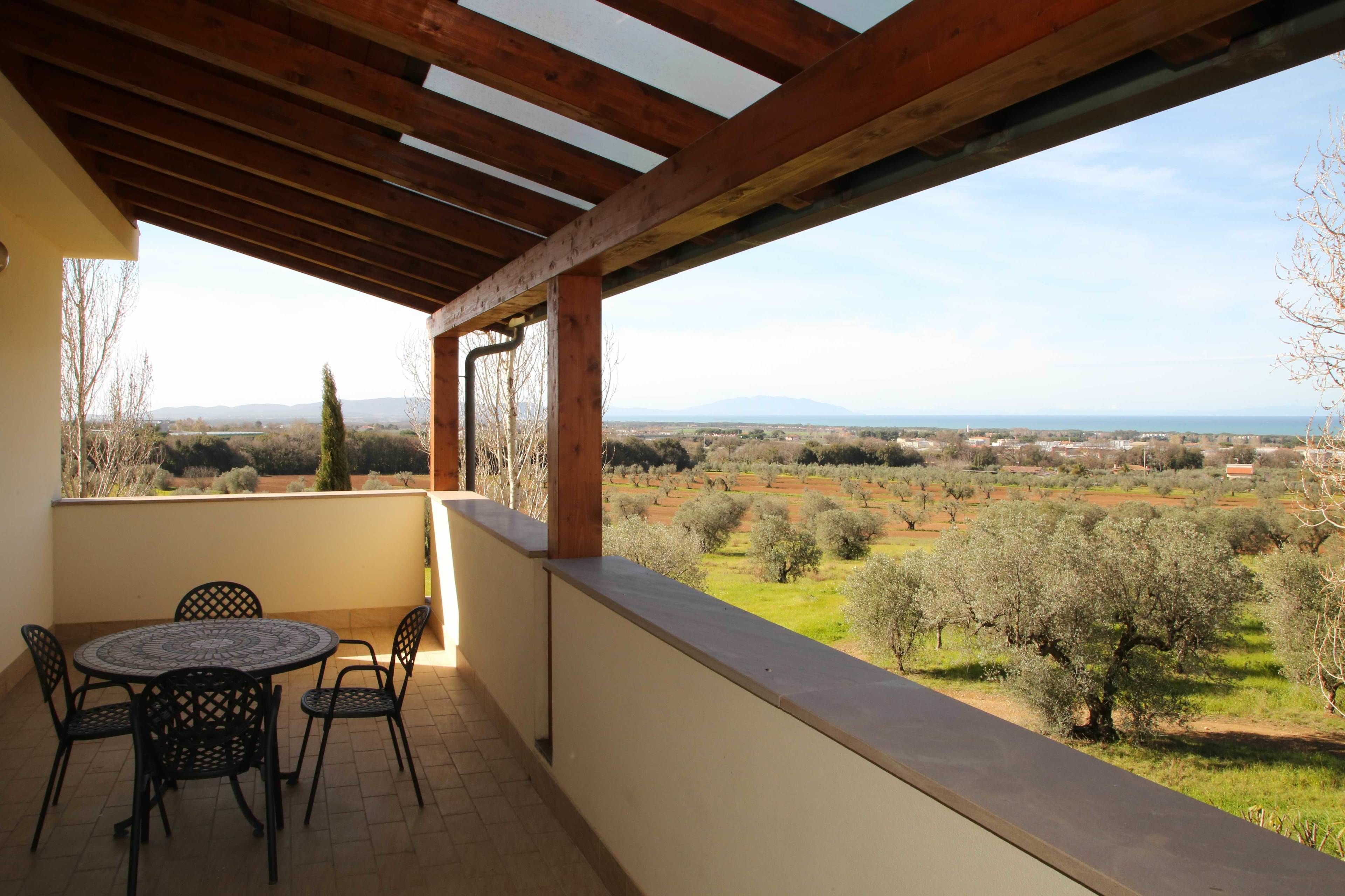 A covered patio with a table and chairs, overlooking a rural landscape with olive trees and distant mountains.