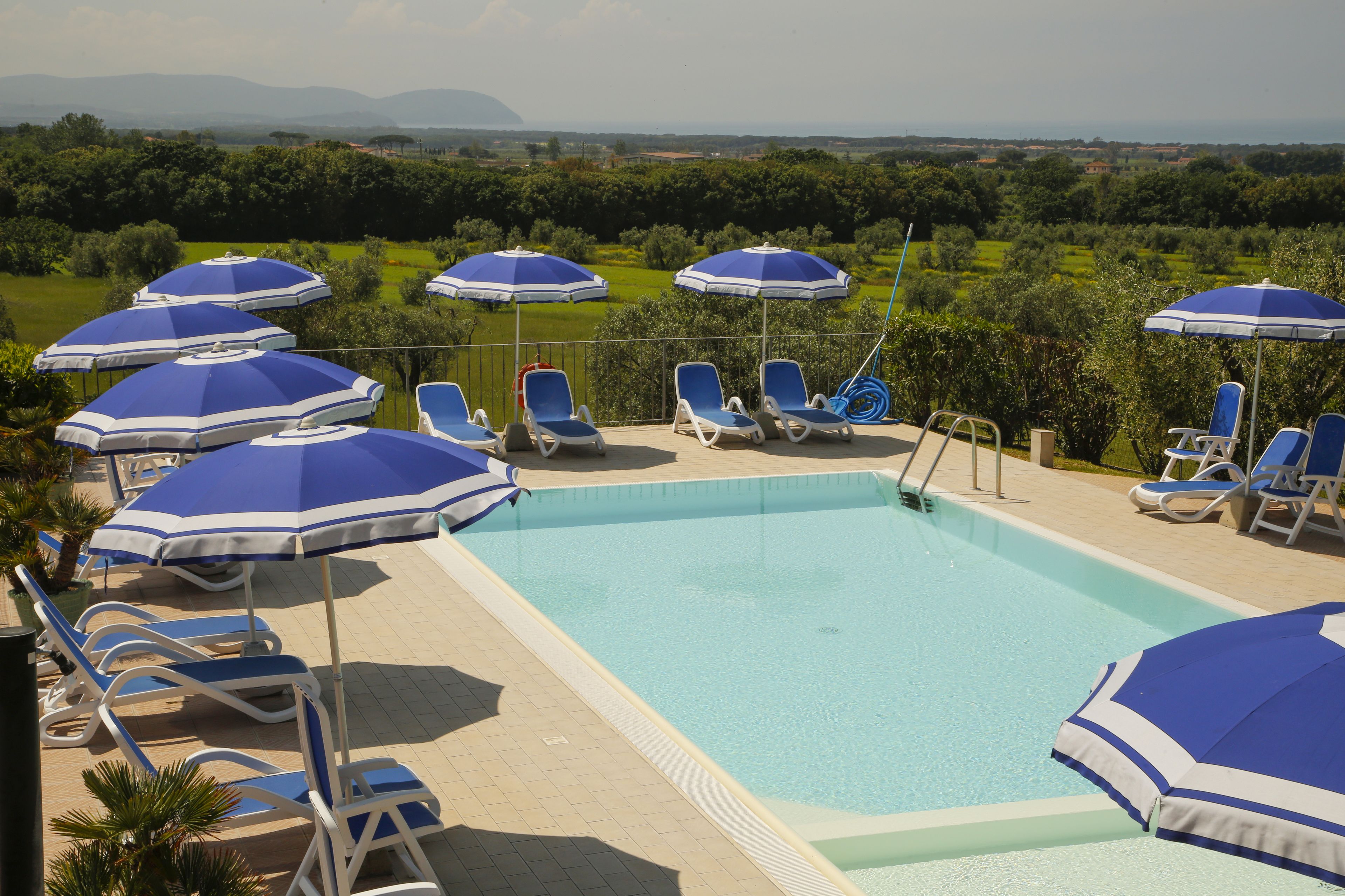 A pool area with blue and white sun umbrellas, lounge chairs, and a view of a green landscape.