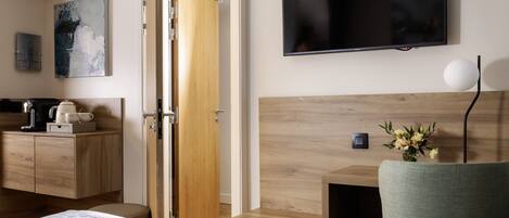 A modern hotel room with a wooden headboard, a flat-screen TV mounted on the wall, a small bedside table with a vase of flowers, and a wooden dresser with a coffee maker.