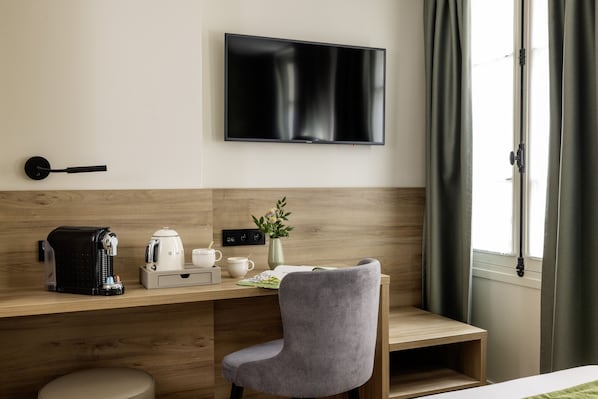 A modern hotel room with a wooden desk, a grey chair, a flat-screen TV mounted on the wall, a coffee maker, a kettle, a vase with flowers, and a window with curtains.