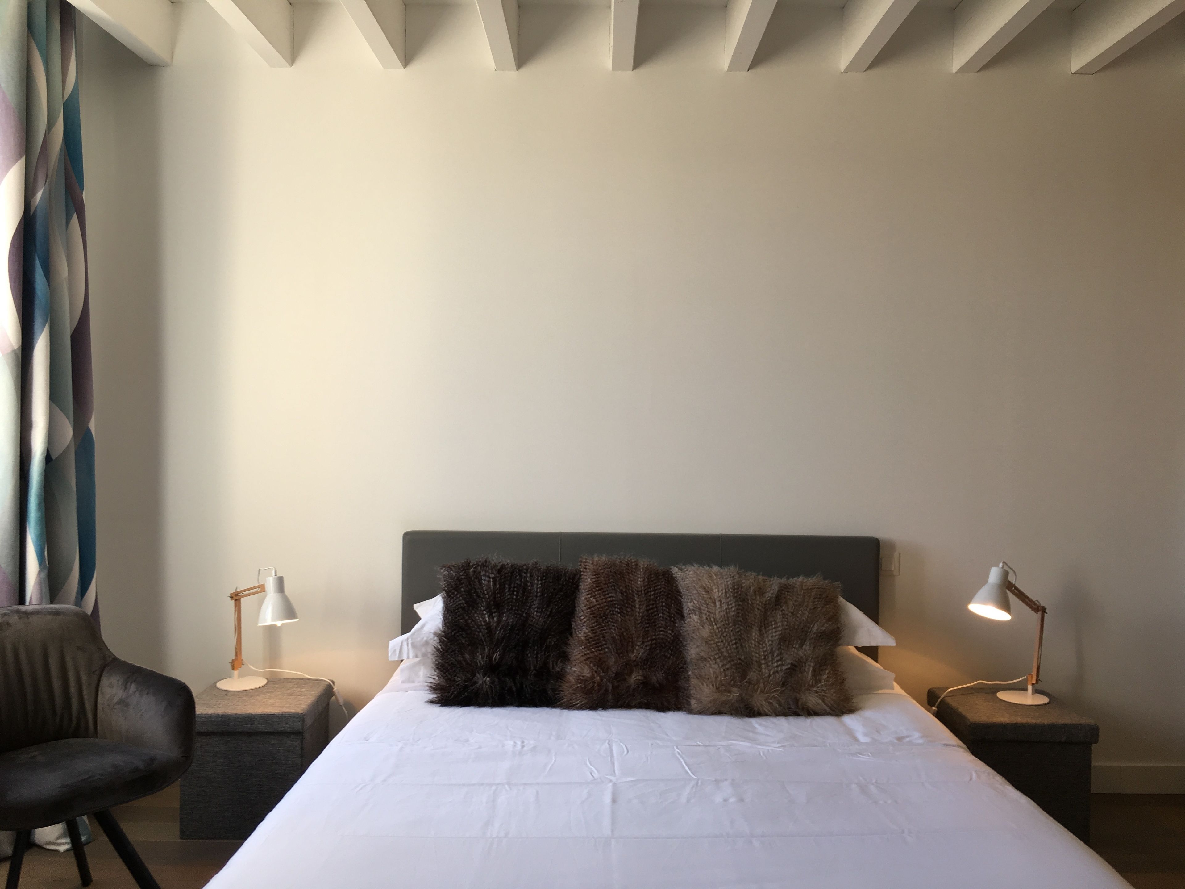 Un lit bien fait, avec une tête de lit sombre, deux tables de chevet avec des lampes, un fauteuil sombre et une fenêtre avec un rideau.