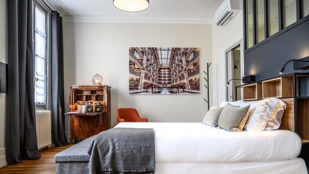 A bedroom with a large bed, a wooden side table, a bookshelf, and a large window with curtains. - Clos 1906 (Saint-Émilion)