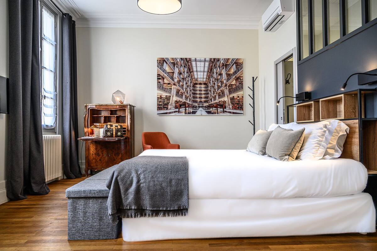 Ein Schlafzimmer mit einem großen Bett, einem Nachttisch aus Holz, einem Bücherregal und einem großen Fenster mit Vorhängen.