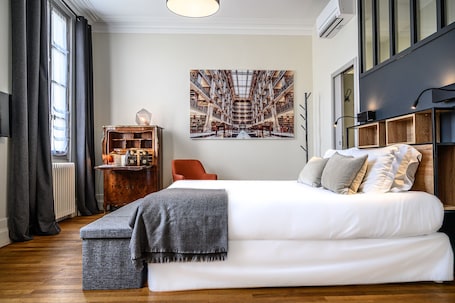 A bedroom with a large bed, a wooden side table, a bookshelf, and a large window with curtains.
