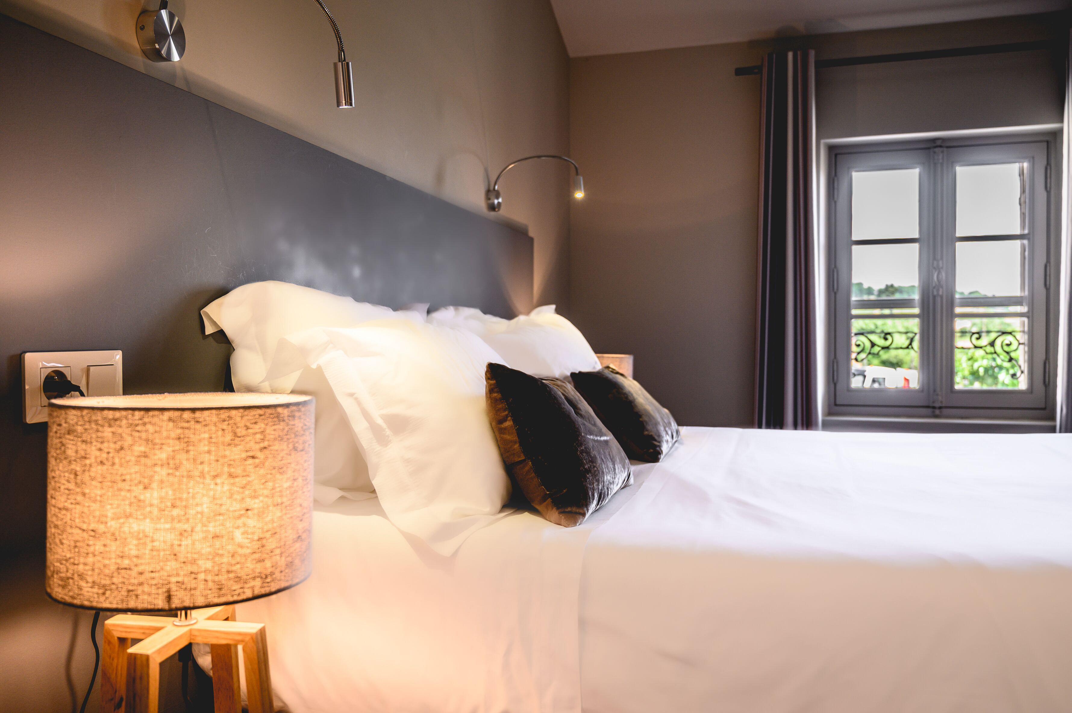 A neatly made bed with white linens and two pillows, a bedside lamp, and a window with a view of greenery.