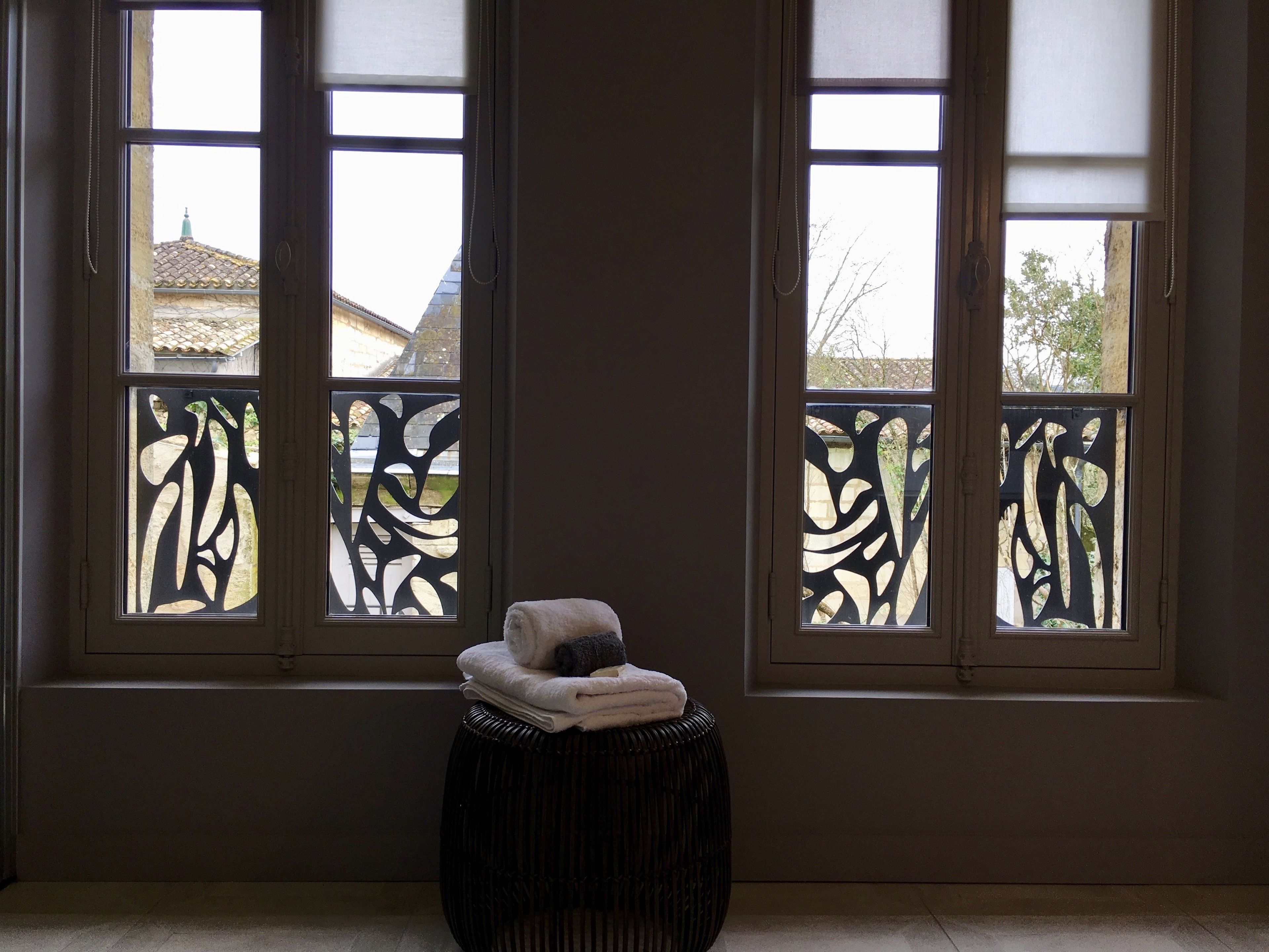 A room with three windows featuring decorative black and white patterns, a small round stool with folded towels, and a view of buildings outside.