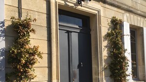 A building facade with a large, dark-colored door, a light fixture above it, and two potted plants on the ground. - Clos 1906 (Saint-Émilion)