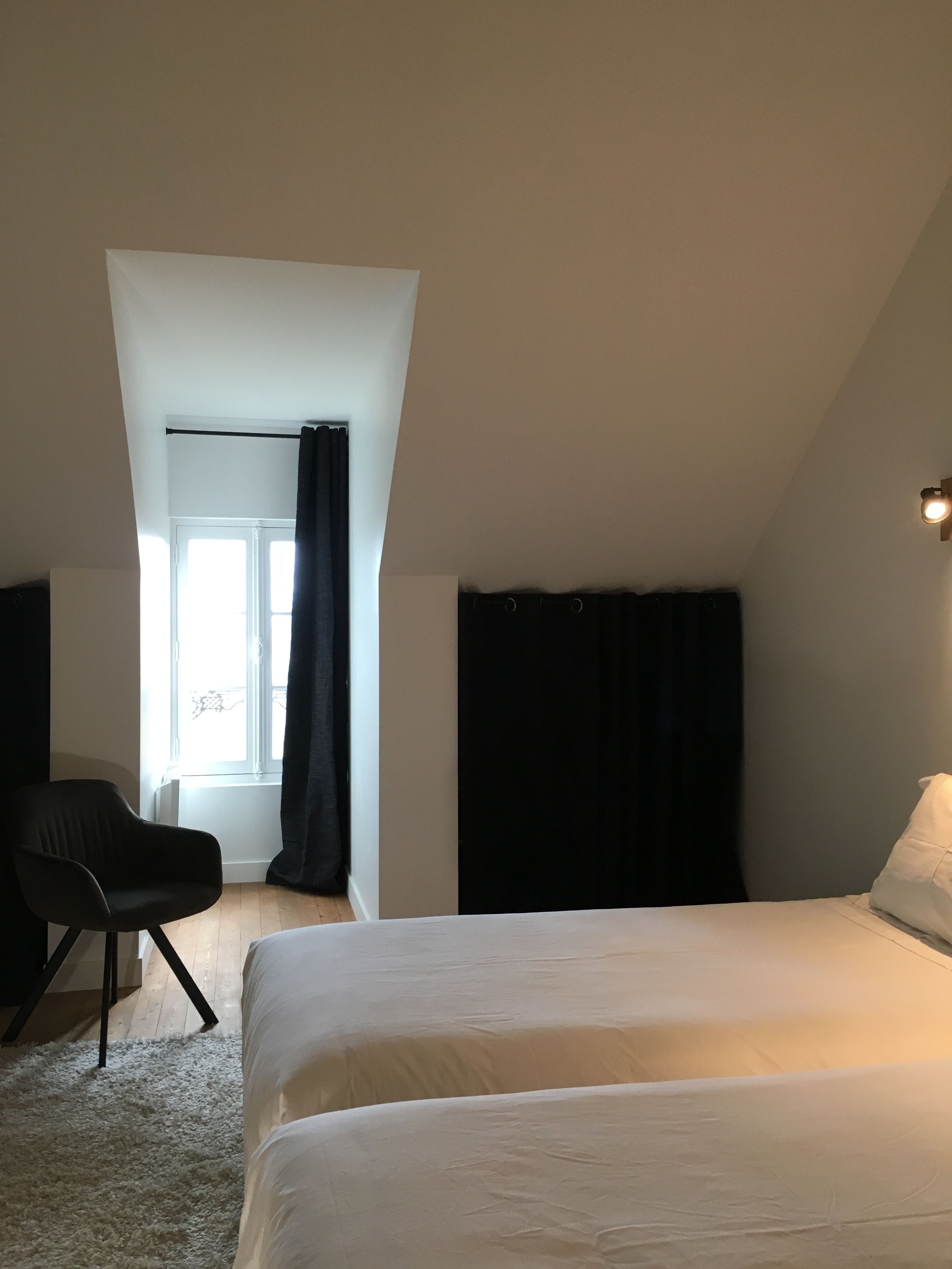 Ein Schlafzimmer mit Bett, Sessel, einem Fenster mit Vorhängen und einer Lampe.