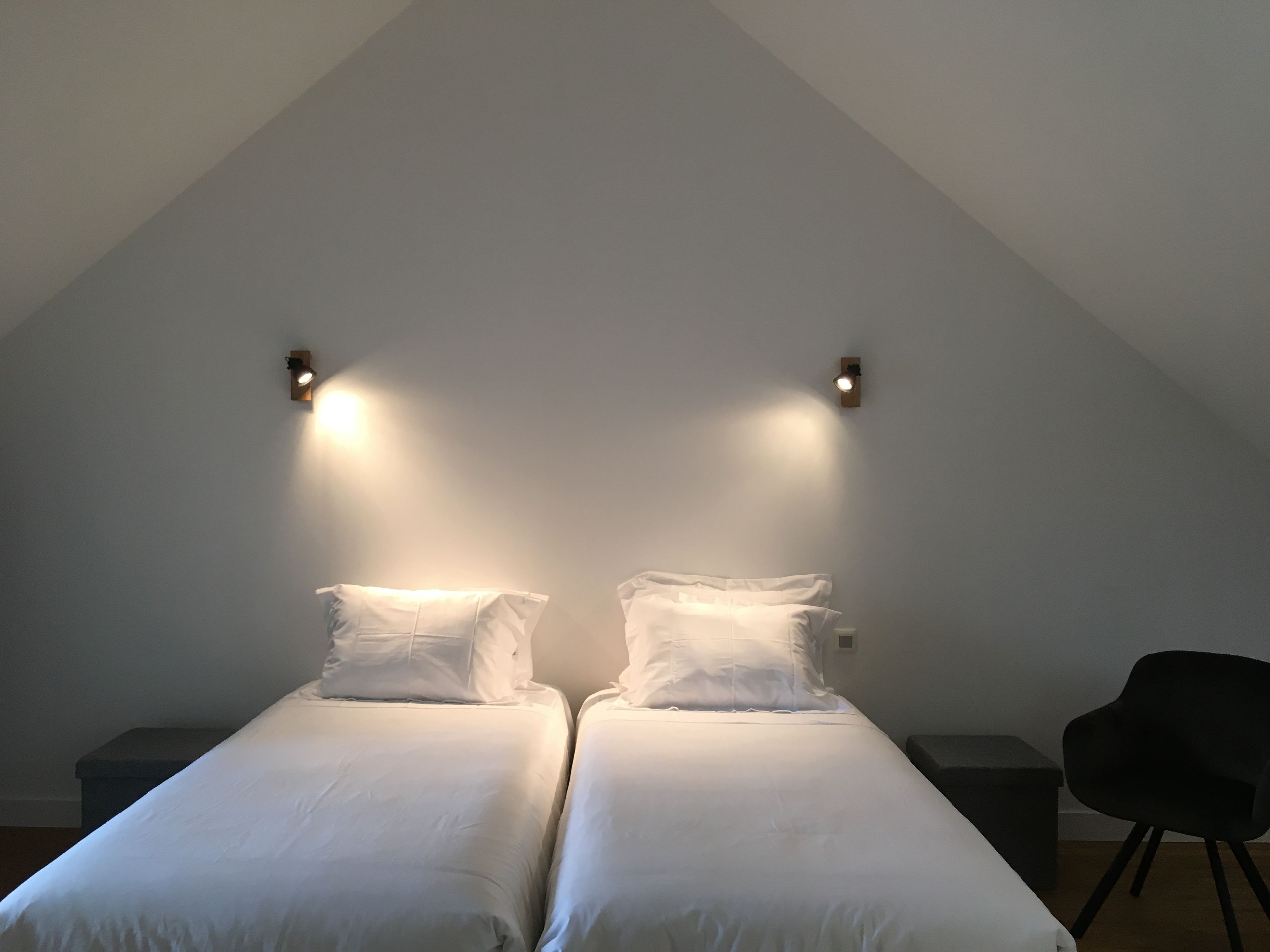 Une chambre avec deux lits, une chaise noire et deux lampes fixées au mur.