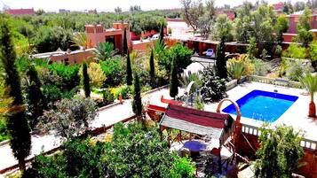 Zona de piscina con piscina azul, rodeada de abundante vegetación y varios edificios.