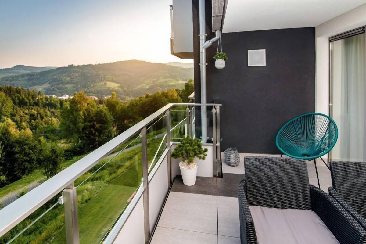 Un balcon donnant sur un paysage verdoyant, un canapé en osier, un fauteuil turquoise et une plante en pot.