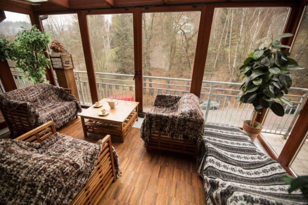 Une véranda avec un plancher en bois, des meubles en osier et une vue sur les arbres à travers de grandes portes vitrées.