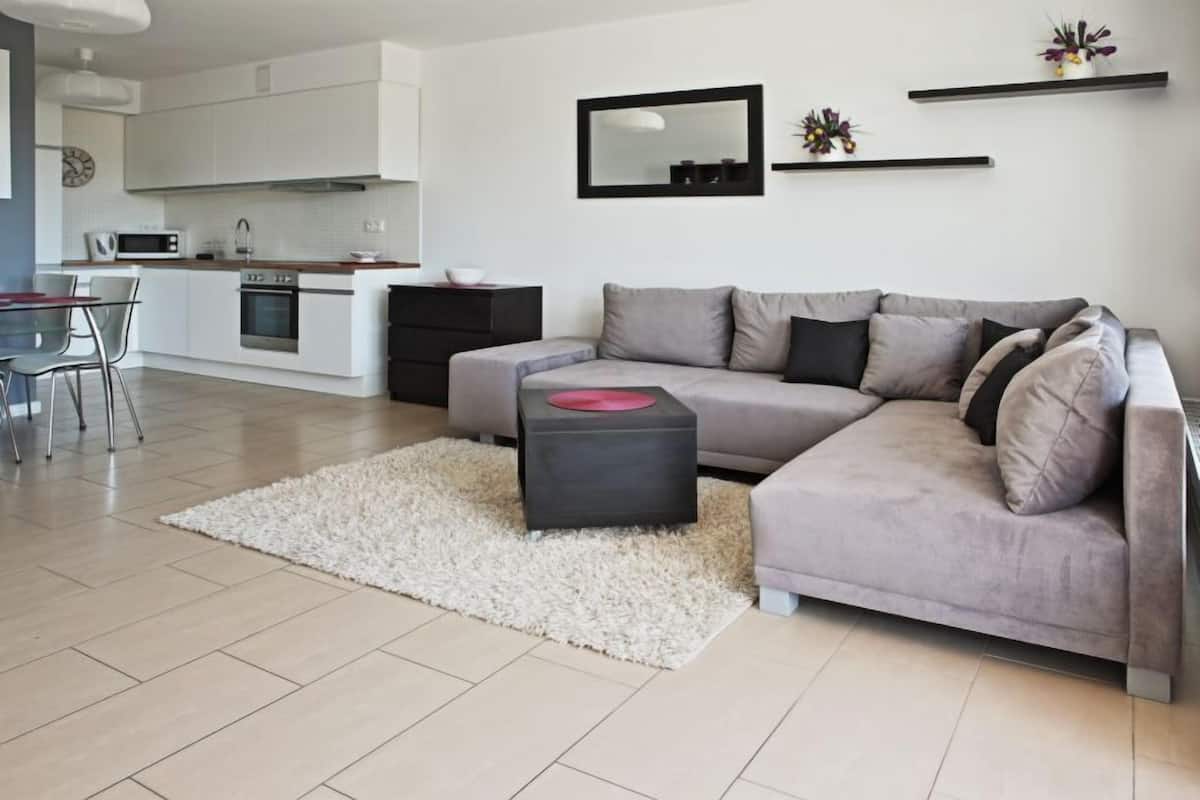 Un salon moderne avec un canapé gris, un petit pouf noir, un tapis blanc et une cuisine en arrière-plan.