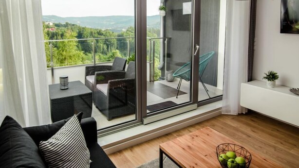 Una sala moderna con un sofá negro, una mesa de centro de madera y un ventanal corredero de vidrio que da a un balcón con muebles de mimbre y vistas al verdor.