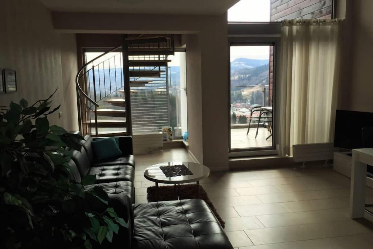 Un salon moderne avec un canapé en cuir noir, une petite table basse et une grande fenêtre donnant sur un balcon et offrant une vue sur la ville.