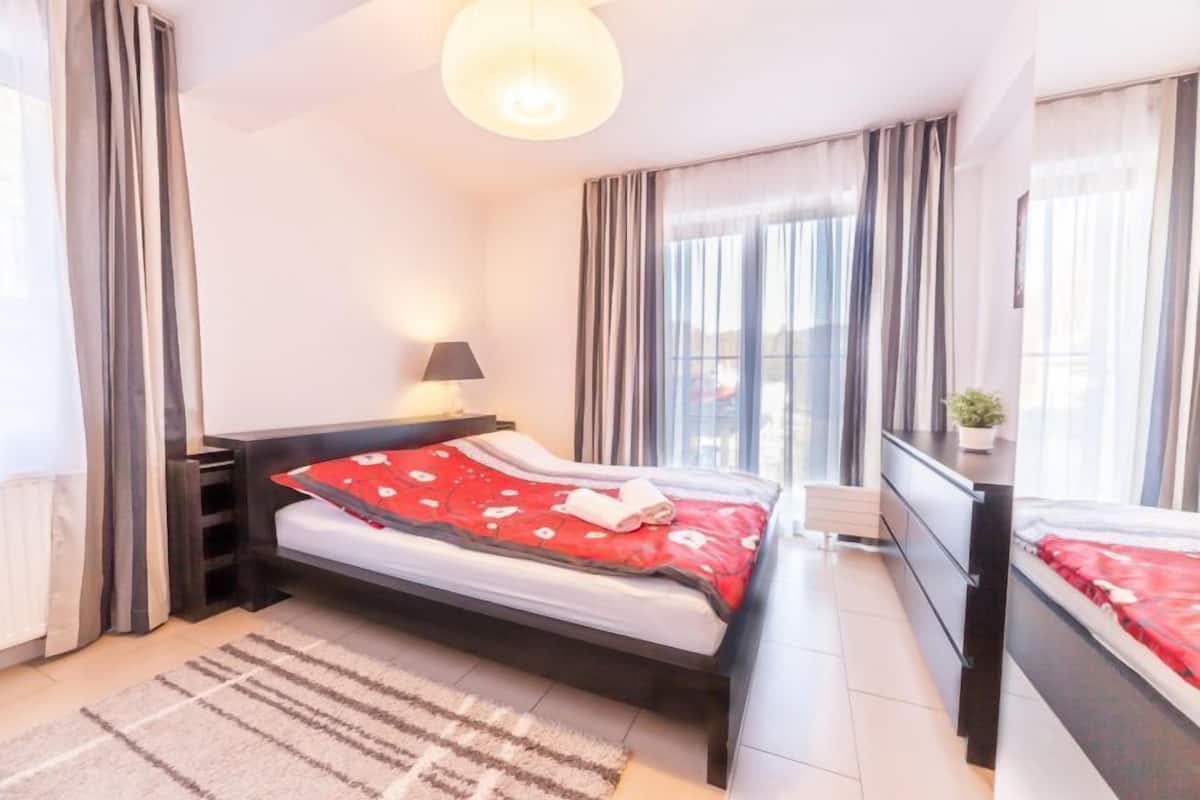 Une chambre moderne avec un lit, des tables de chevet, une lampe et une vue sur la ville grâce à de grandes fenêtres.