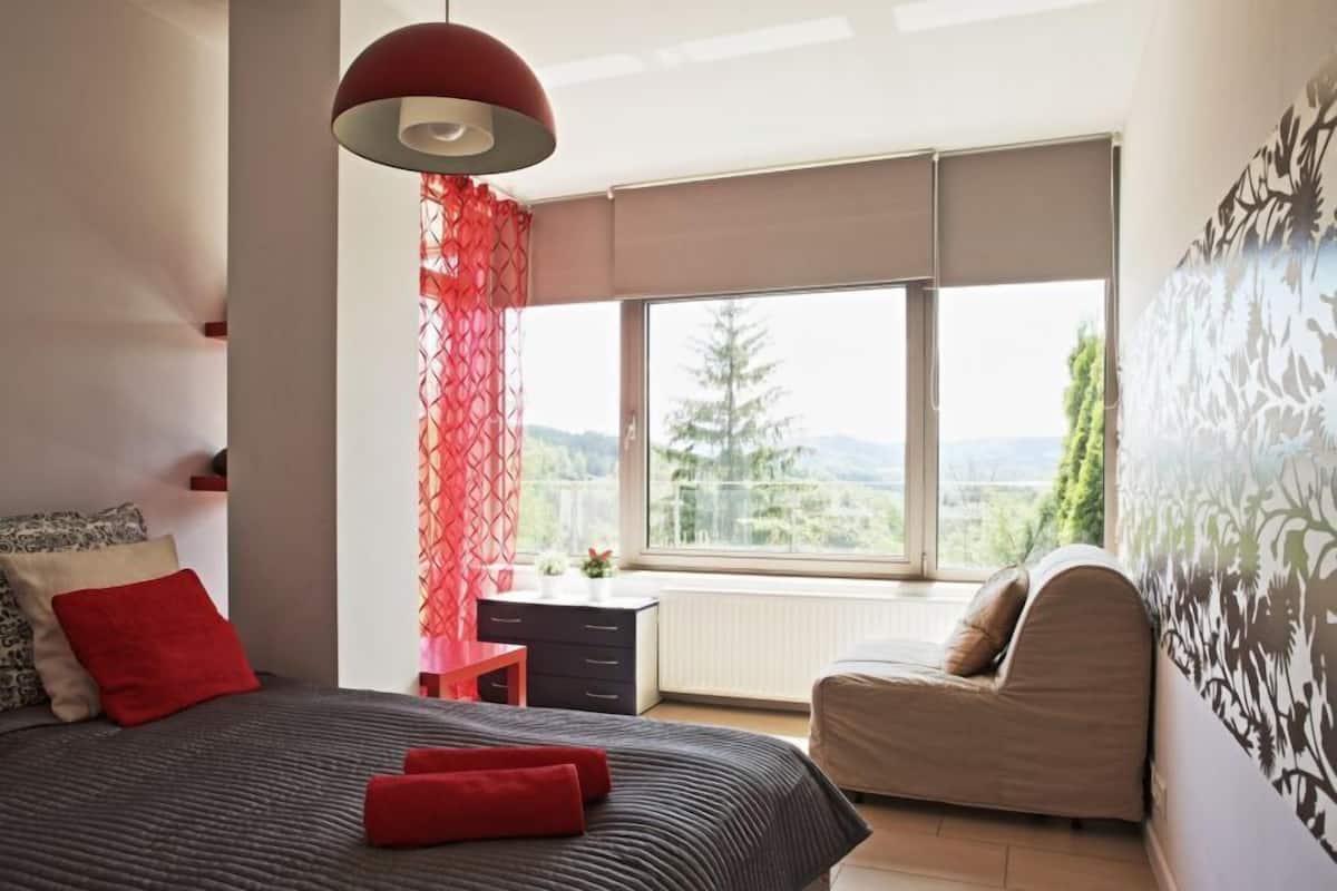 Une chambre moderne avec un grand lit, un bureau et une chaise. Une fenêtre offre une vue sur les arbres et les montagnes.