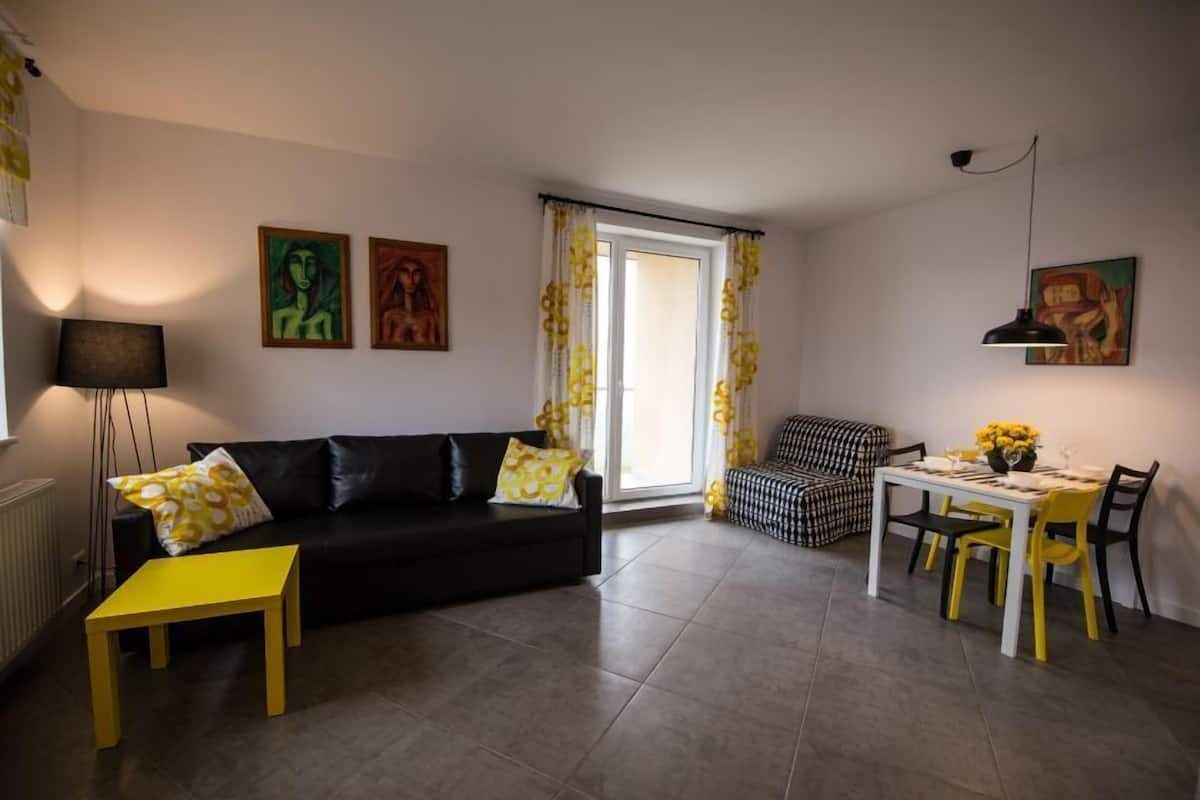 Un salon avec un canapé noir, une table jaune et un coin repas avec une nappe à carreaux.