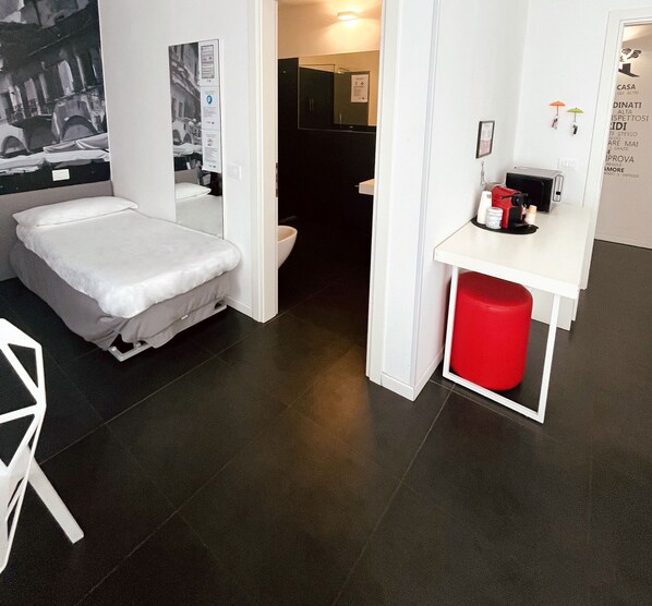 A compact hotel room with a bed, a small desk with a coffee machine, a red trash bin, and a bathroom visible through an open door. - DiMoras House (Verona)
