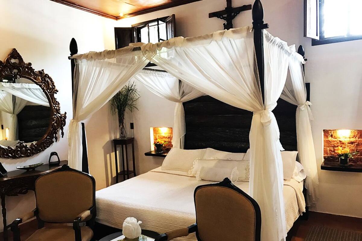 A four-poster canopy bed with a headboard, two chairs, a bedside table with a lamp, and a mirror.